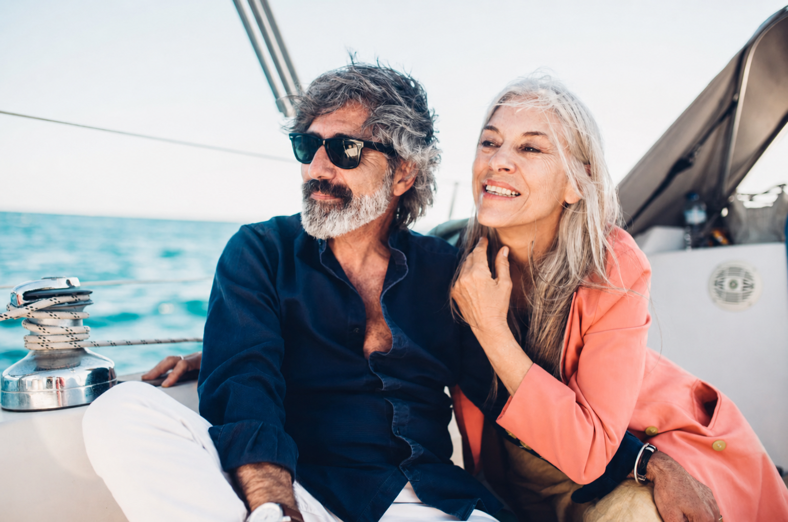 An older man with gray hair and beard, wearing sunglasses and a navy blue shirt, sits beside a smiling older woman with long gray hair in an orange jacket; they are on a boat with the ocean in the background, enjoying a sunny day.