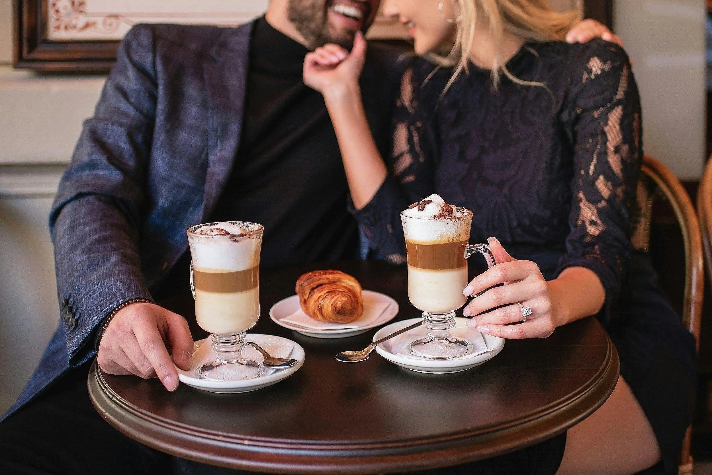 A couple sitting at a table enjoying two frappuccino coffee drinks with whipped cream, chocolate drizzle, and cocoa powder, along with a croissant placed on a small plate. They are smiling and leaning in close to each other.