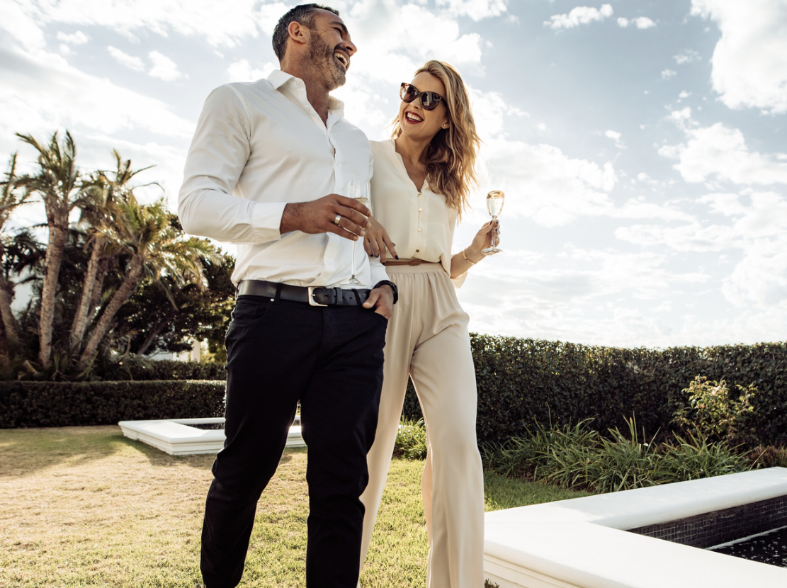 A man and woman outdoors, smiling and holding glasses of wine, with palm trees and a hedge in the background.