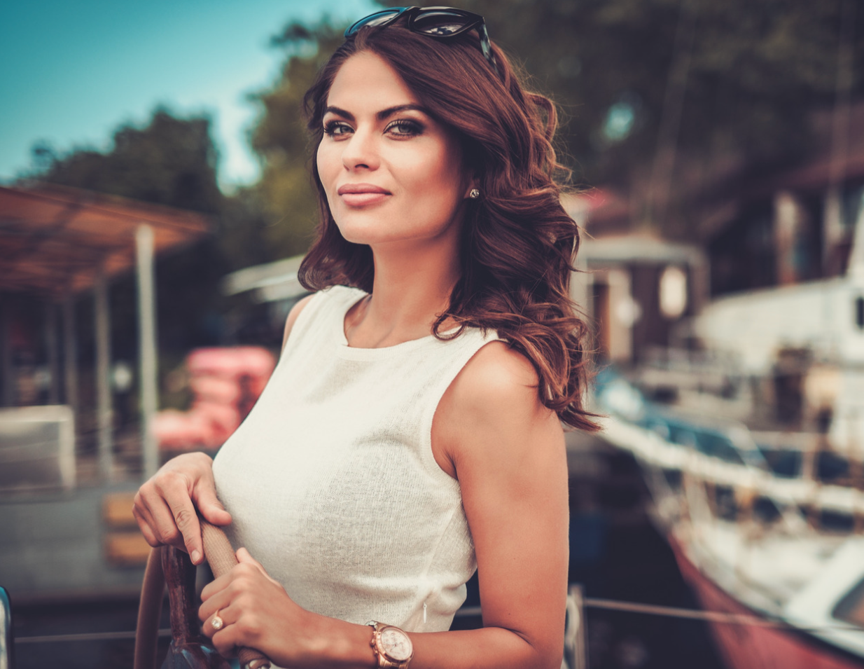 A woman with wavy brown hair wearing a sleeveless white top and sunglasses on her head, standing outdoors near boats or yachts.