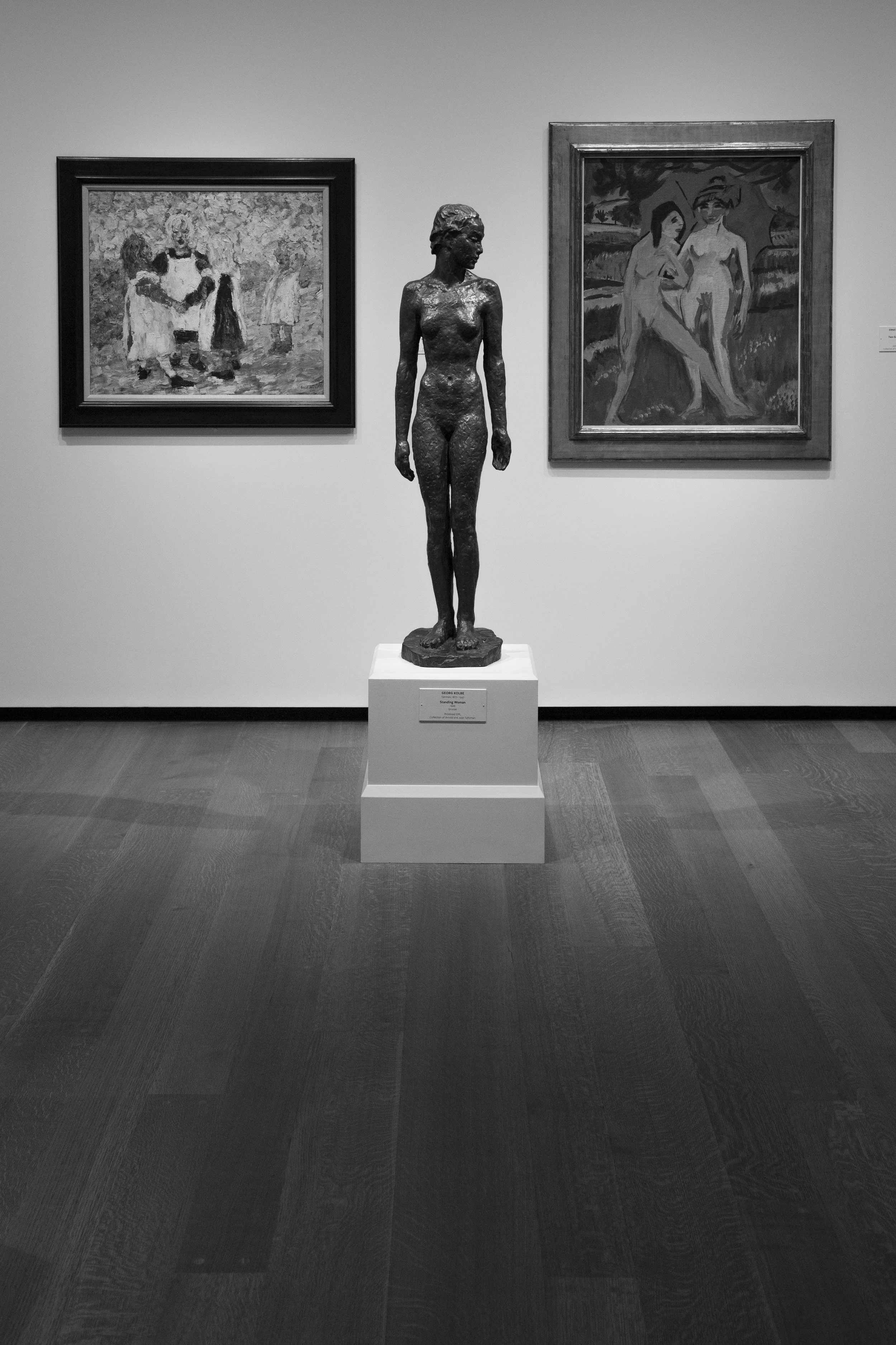 Symmetrical black and white photo of statue and paintings in museum setting, beautiful room with wood flooring. 