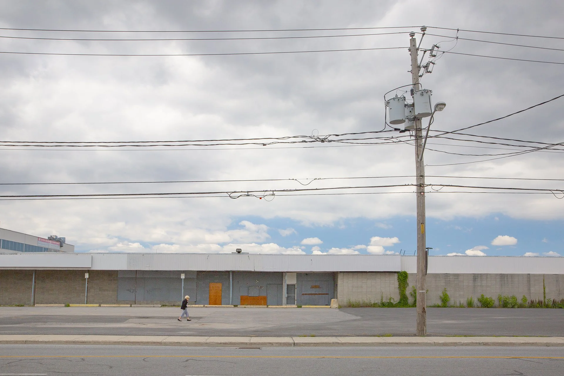 Solitary figure walking past a shuttered strip mall under power lines and a cloudy sky, Montreal. Photo by Lionel May