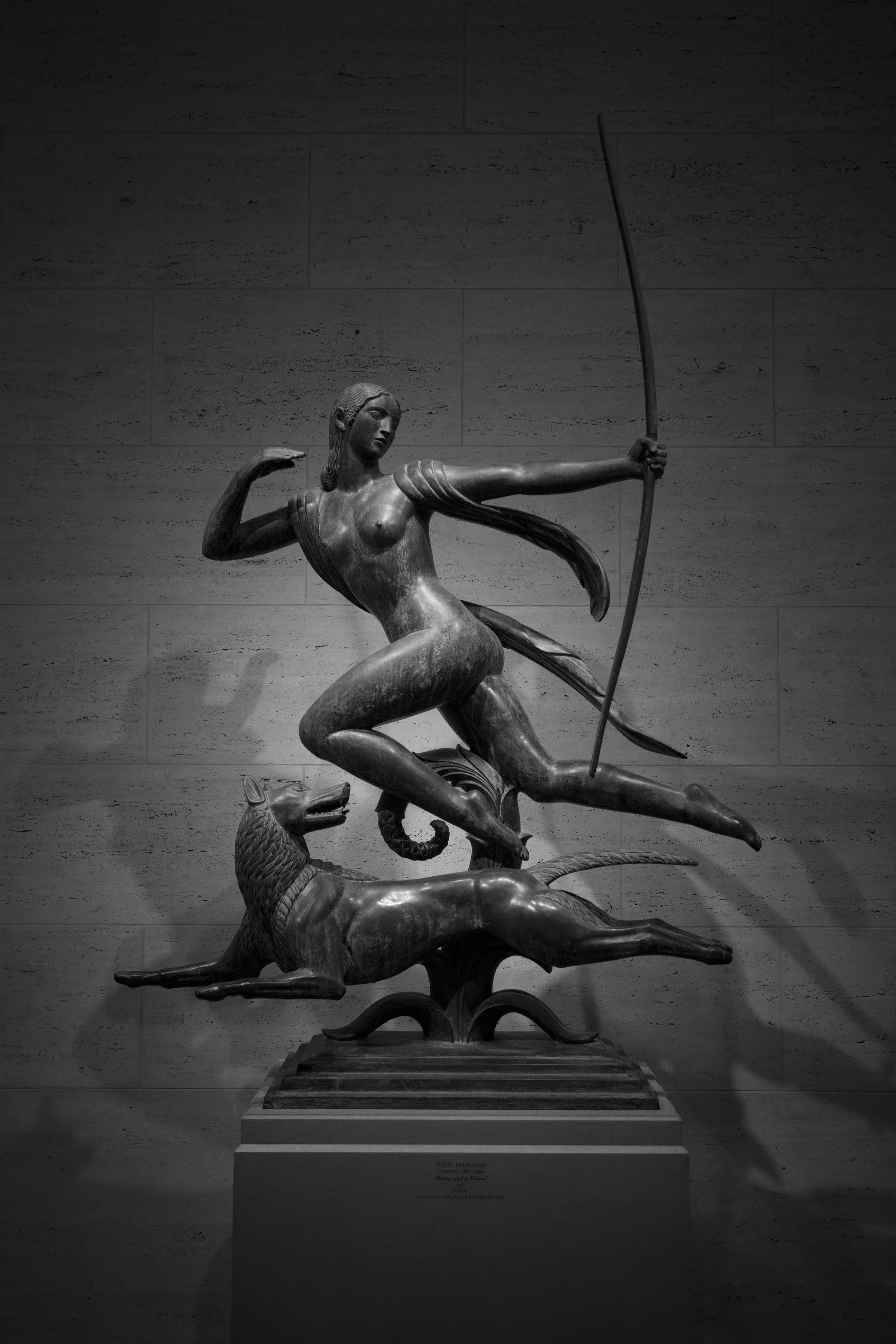 Satue of huntress in mid jump with dog, in art museum, highlighted by focused lighting against wall. 