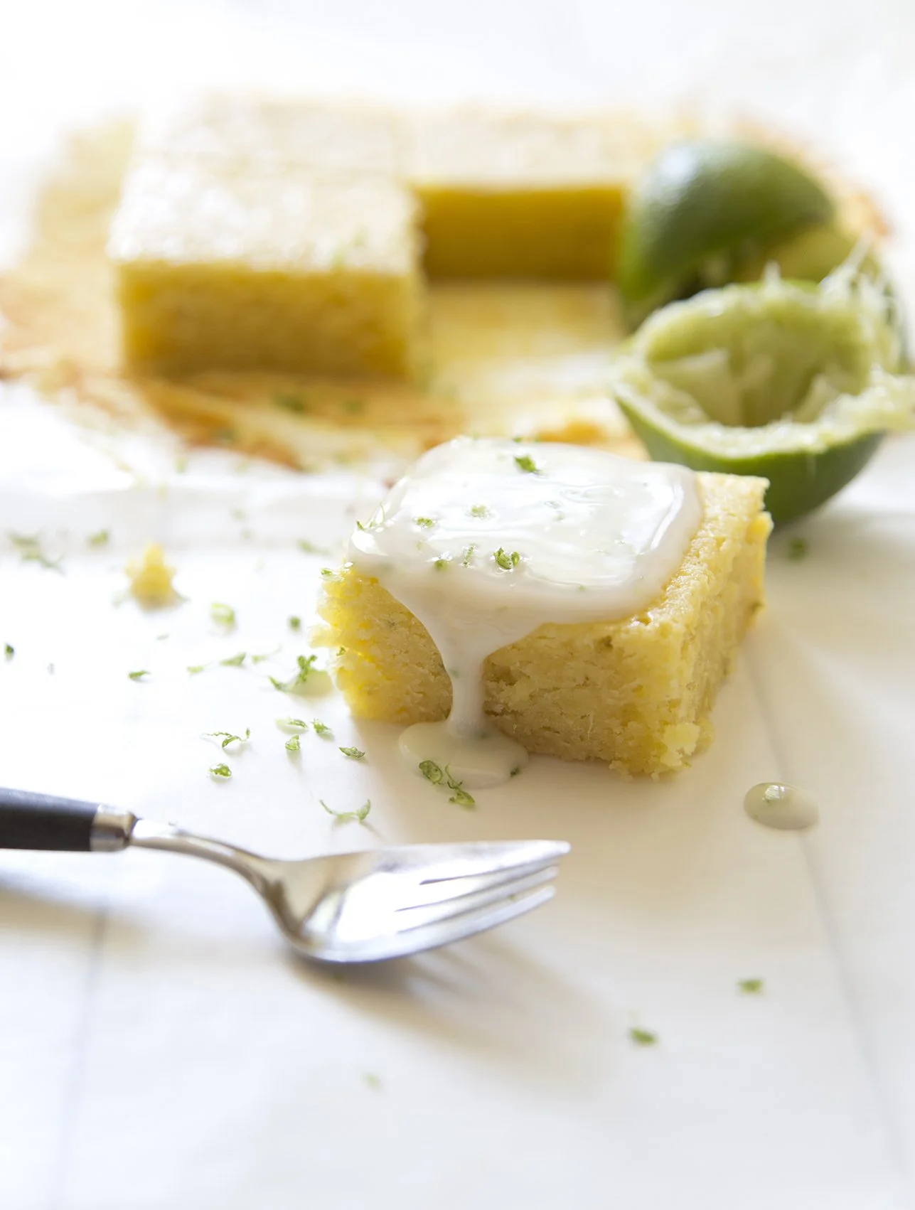 Lime and whhite chocolate brownies, with lime glaze and lime zest on top, on white presentation board and elegant fork. 