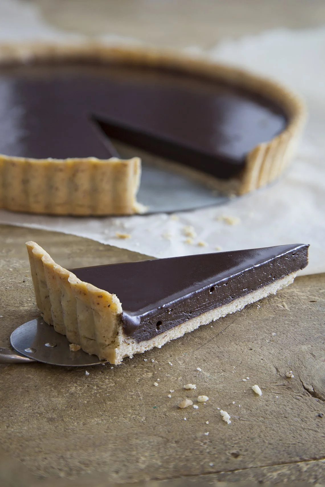 Silky smooth chocolate tart, a thin slice being served, with the rest of the dark chocolate pie visible in soft focu in the background
