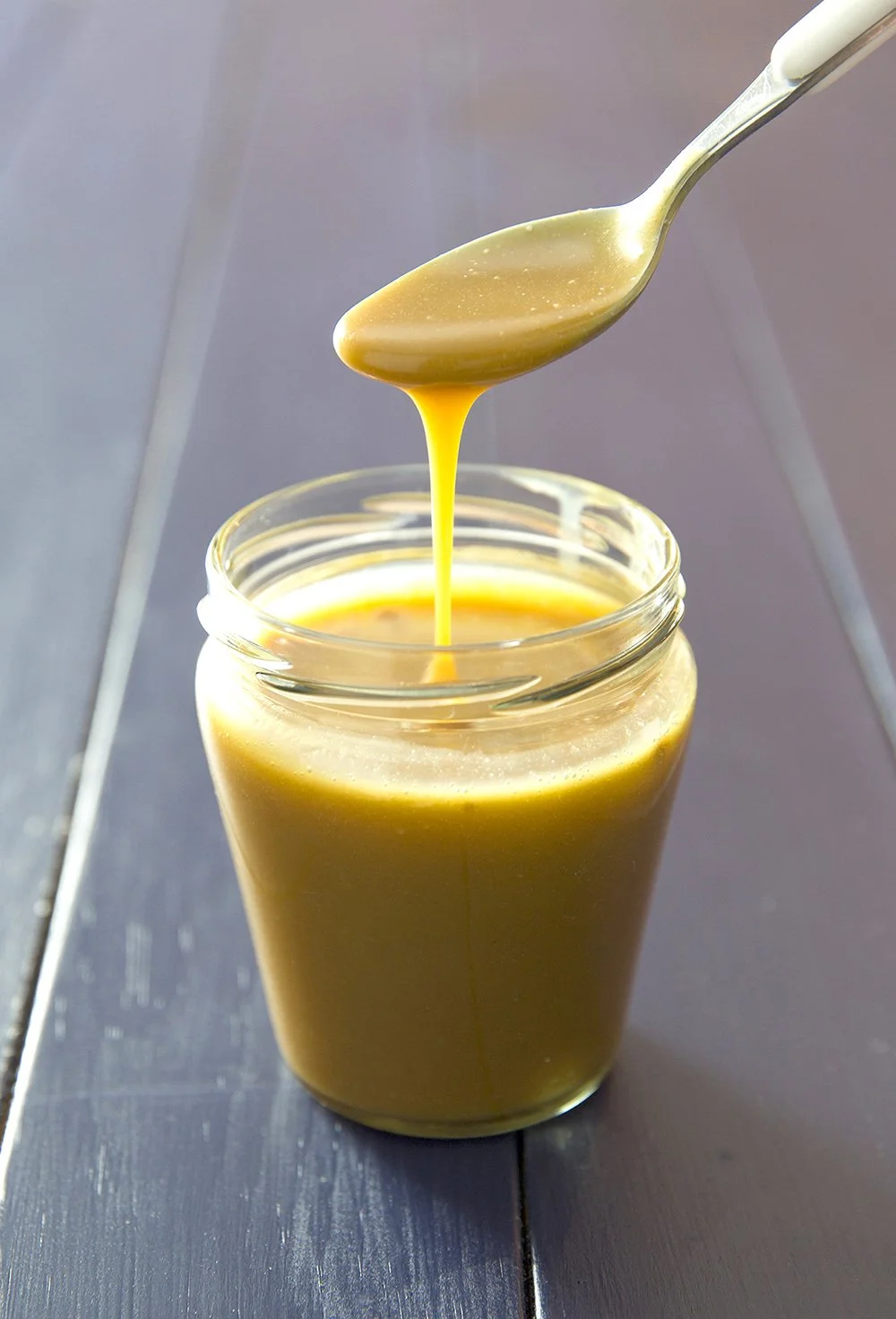 Jar of golden slated caramel with spoon dripping caramel, on blue wooden table