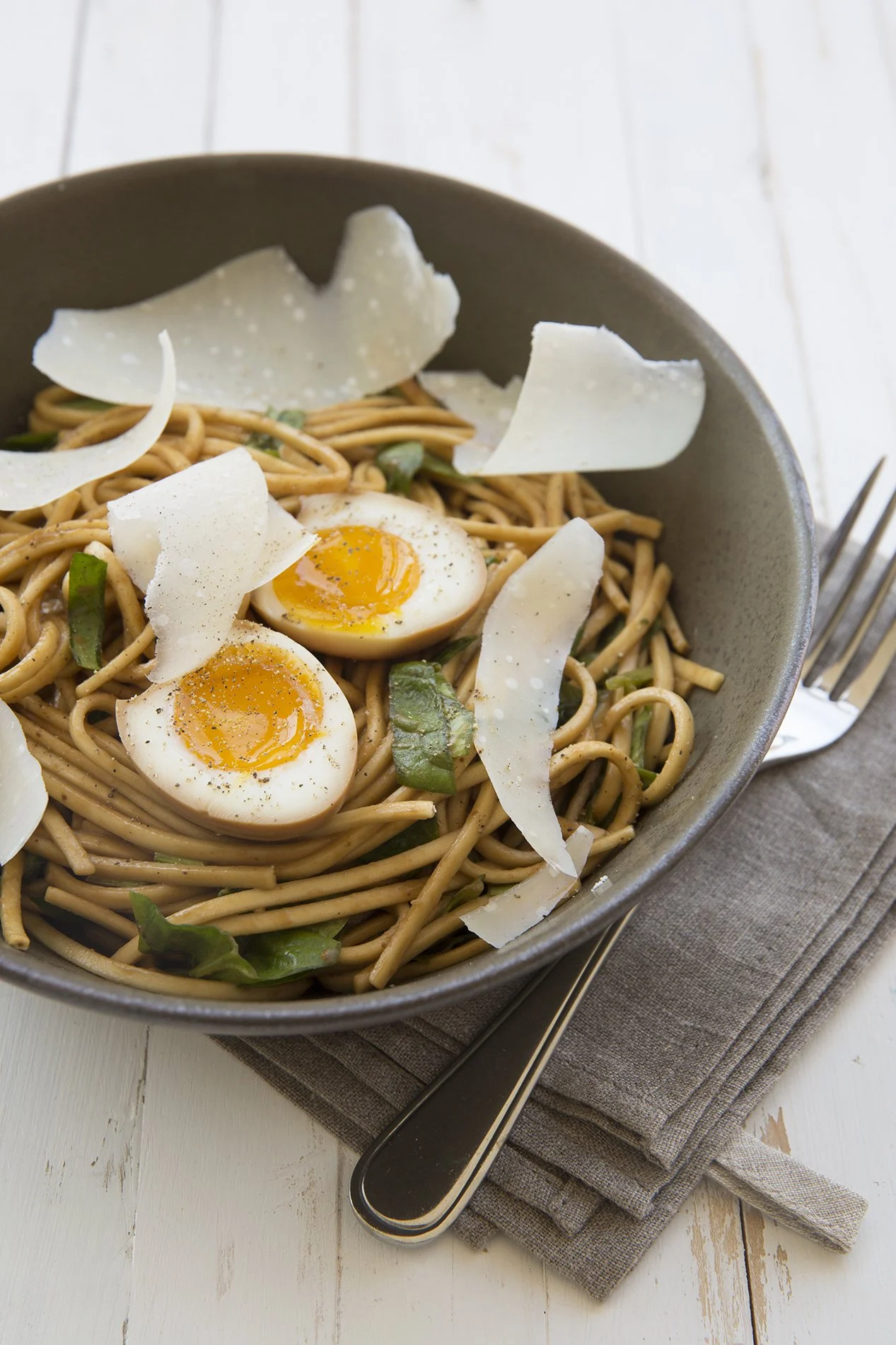 A bowl of japanese noodles with soft boiled eggs and curls of parmesan in a grey bowl on a white wooden table