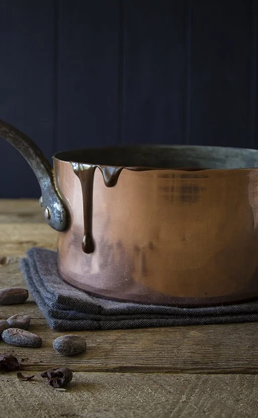 Copper saucepan with melted dark chocolate on rustic wooden board with cocoa beans 