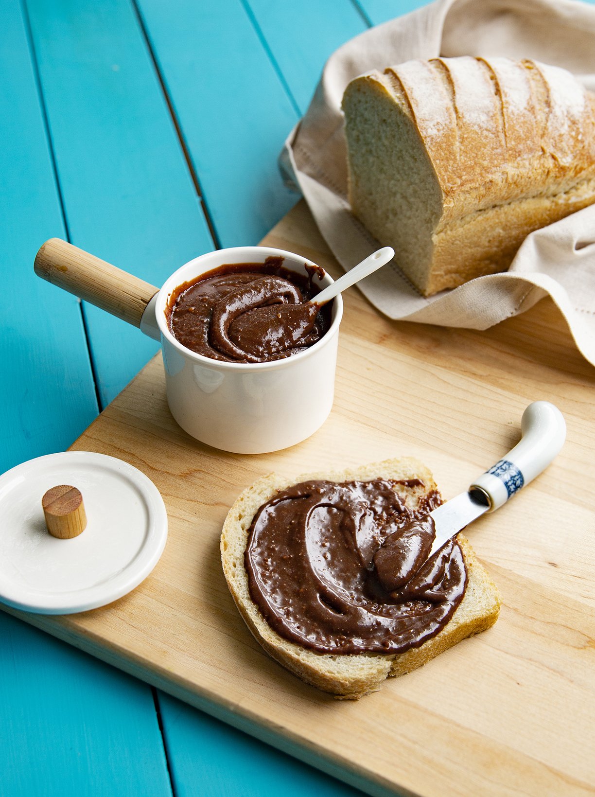 Chocolate and hazelnut spread in a white porcelain serving jar, with a slice of bread and a knife
