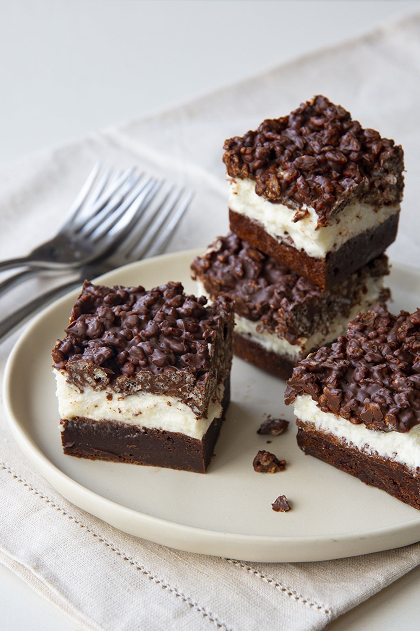 Thick brownies with a chocolate layer, a cream layer and a crunchy topping, arranged on a white plate. 