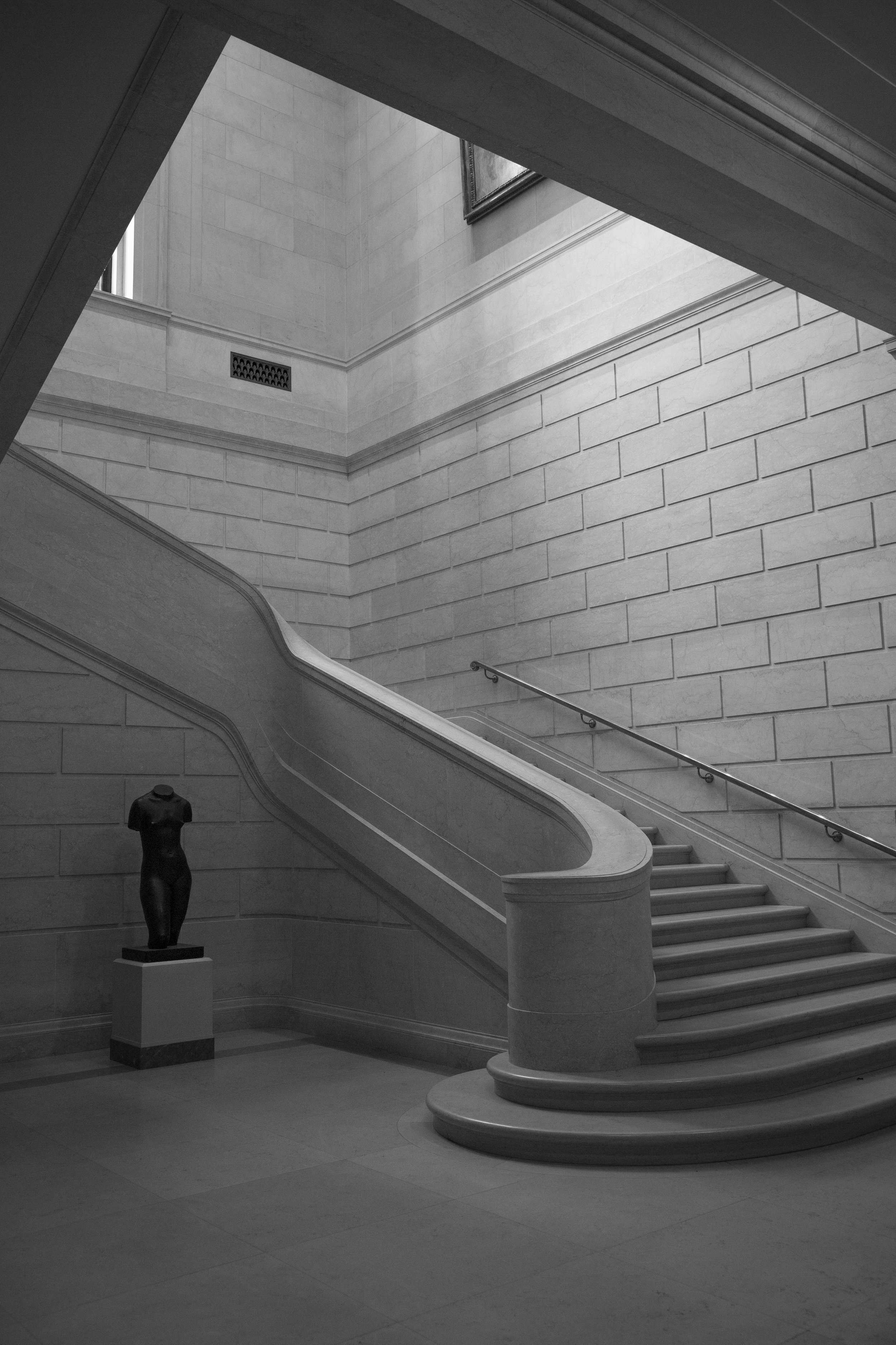 Elegant curved stone staircase in art museum, with sunlight pouring in from the ceiling, and lone statue, dark against the white stone. 