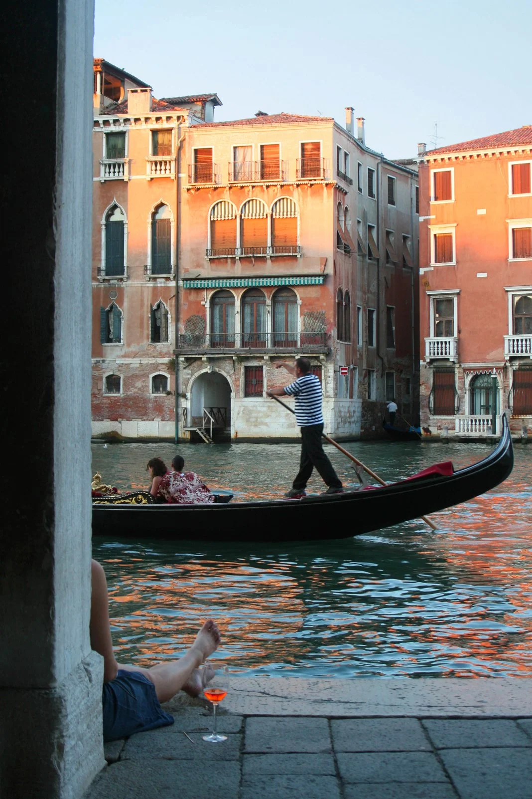 Venice canal, orange and pink ventian buildings with gondola, and people having a drink in front of the canal as the sun sets. 