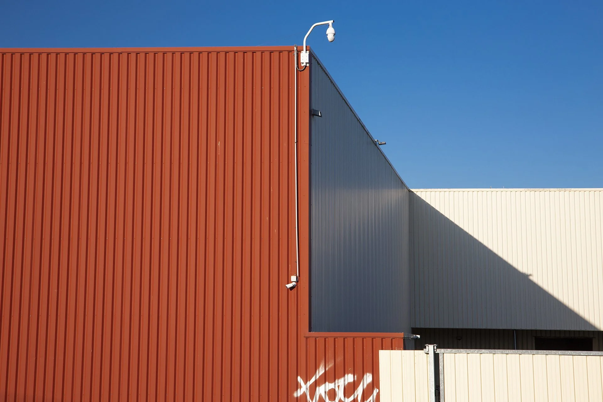 Industrial building facade in rust red, dark grey, and cream corrugated metal panels against a clear blue sky, Sarzeau, France.