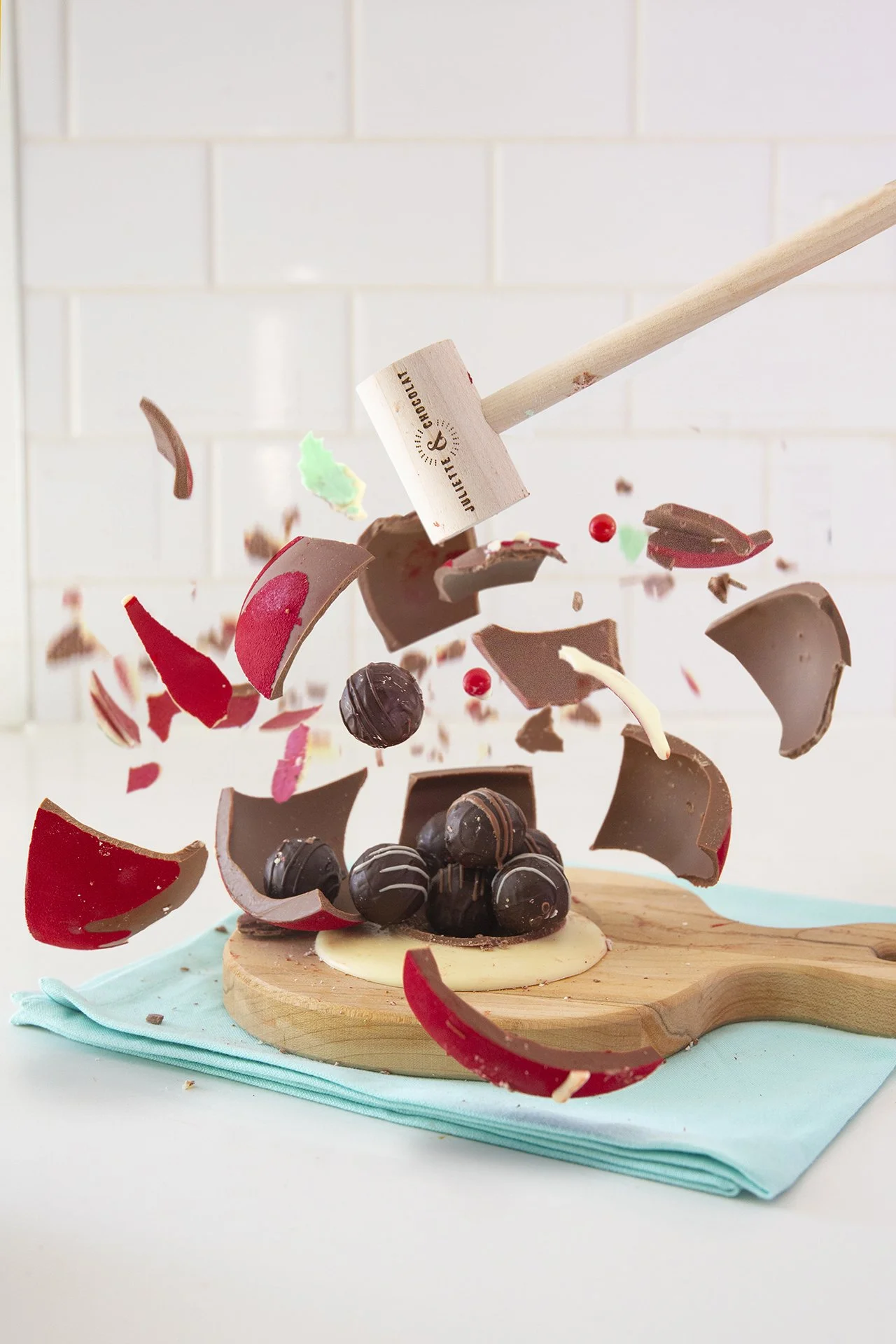 Small wooden hammer smashing a chocolate Christmas ornament, with pieces of chocolate flying everywhere, and chocolate truffles at the center