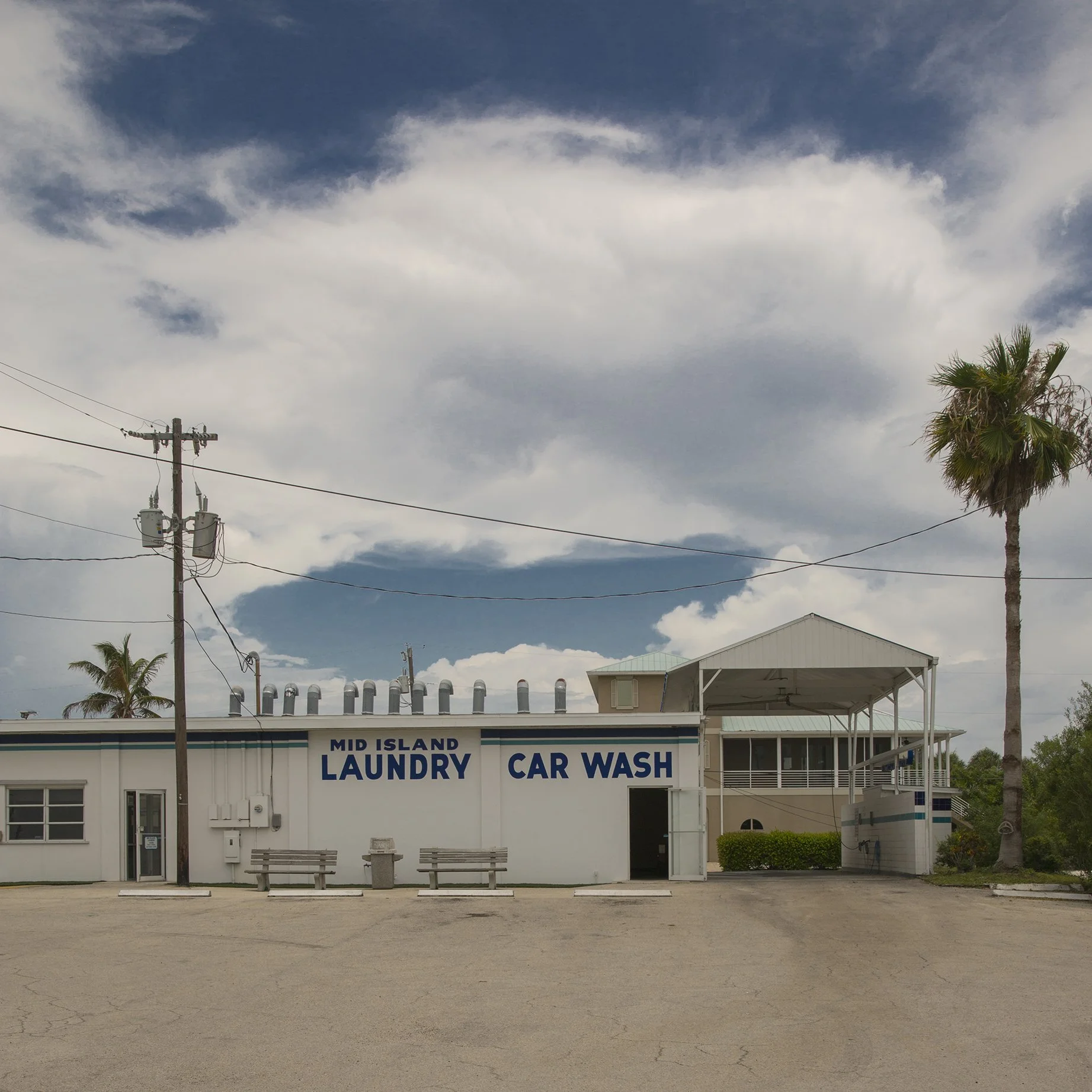 Mid Island Laundry and Car Wash storefront under a large cloud, palm trees and power lines, Florida. Part of Lionel May's photography series on suburban vernacular architecture.
