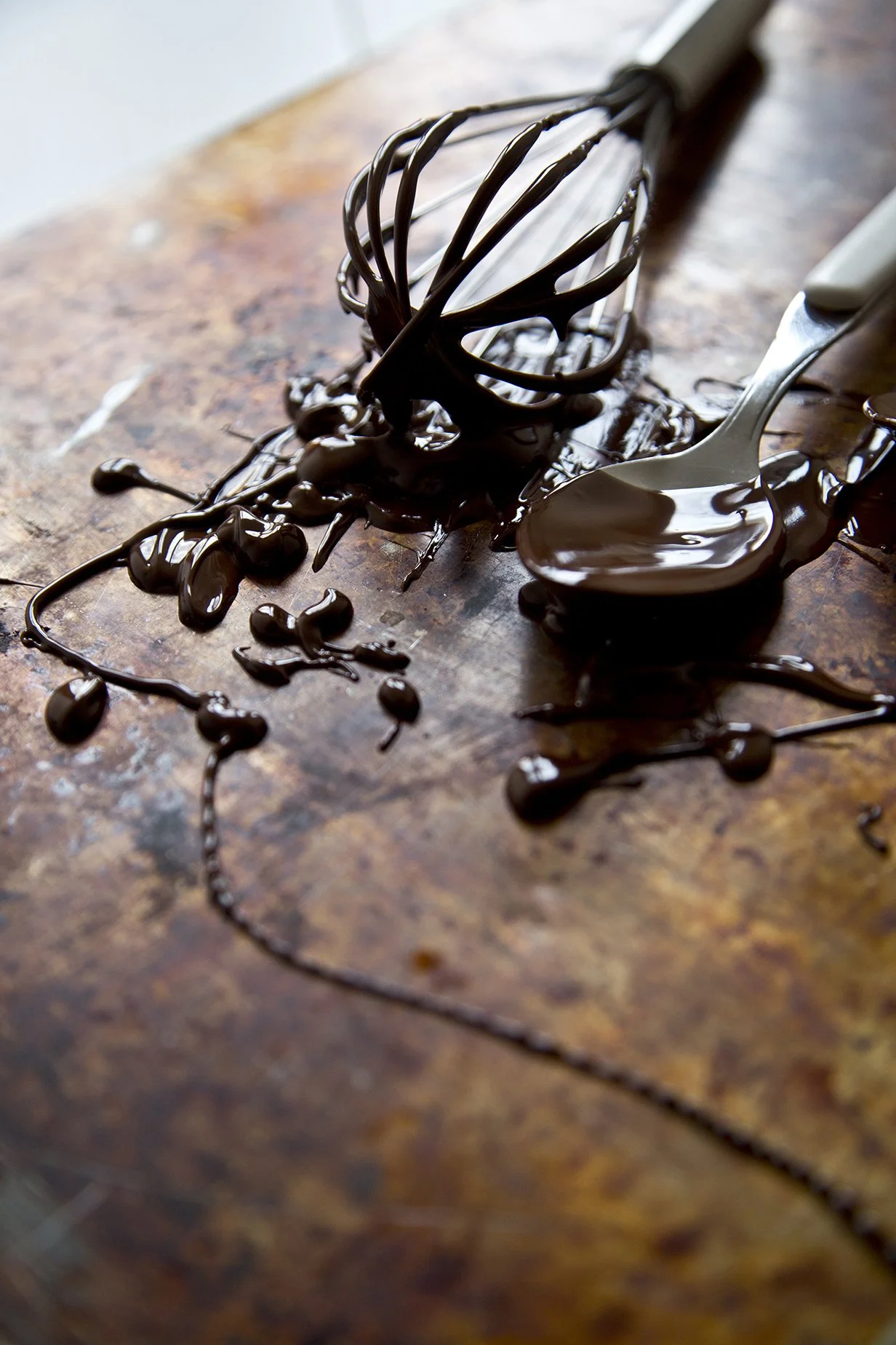 Melted chocolate dripping off a whisk and spoons on a rustic metal surface.
