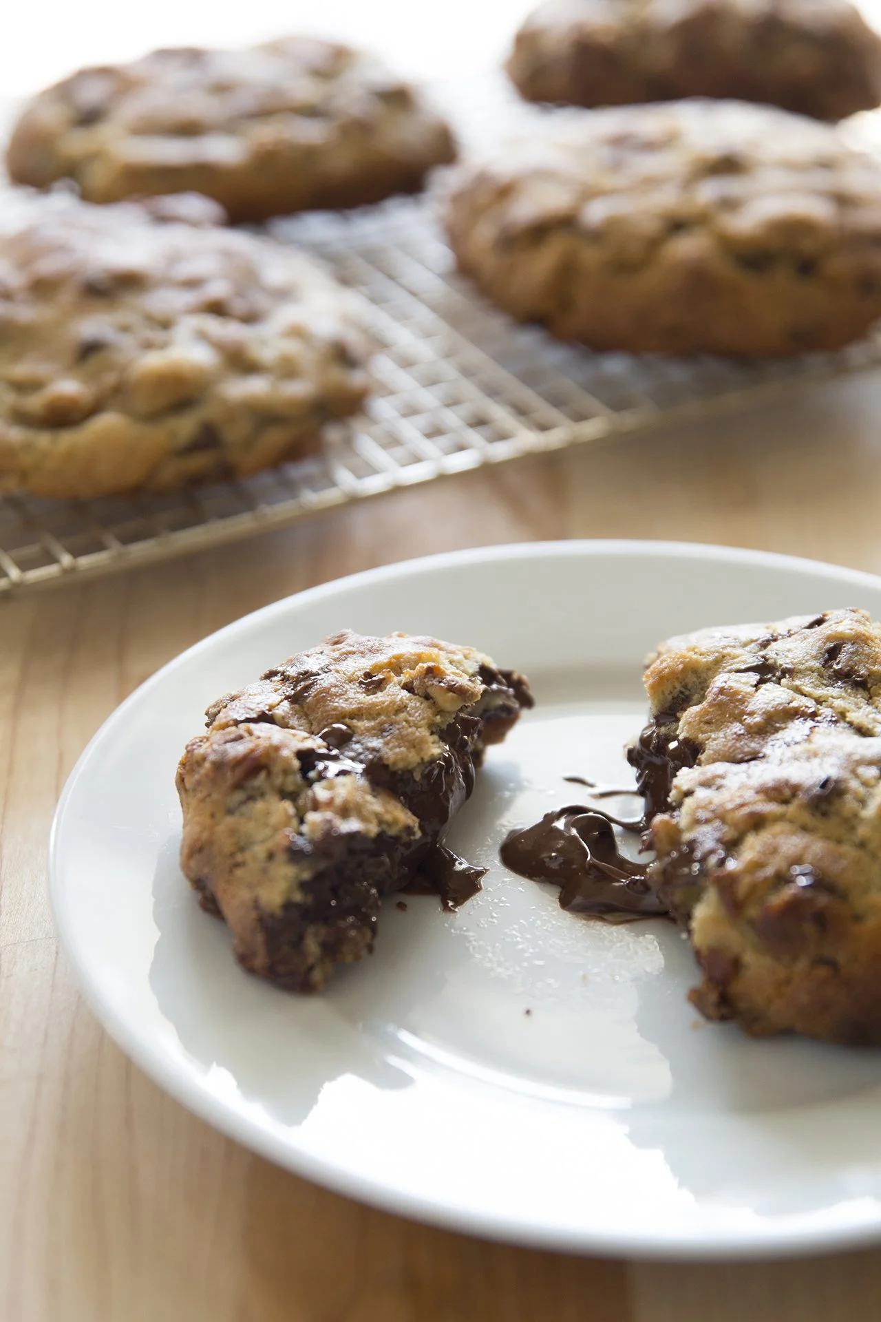 Large chocolate chip cookies with molten praline heart, one cookie split open on a white plate, and more cookies on a cooling rack in the background
