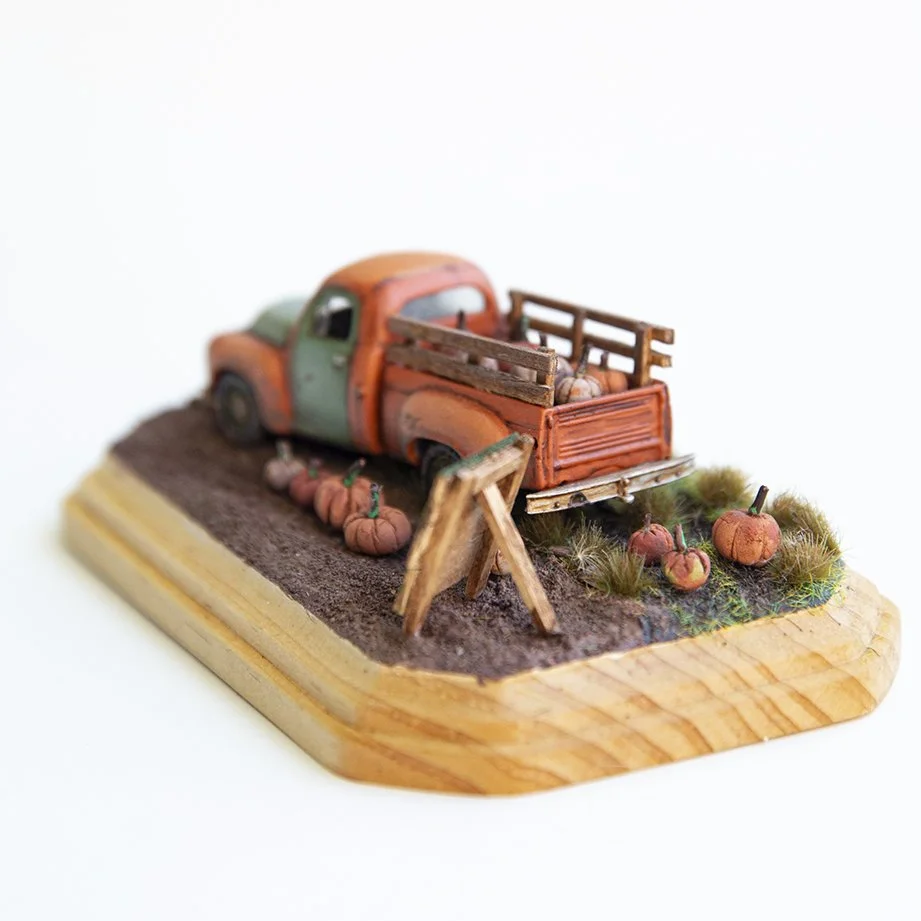 A scale model miniature pick-up truck carrying pumpkins, on diorama base. 