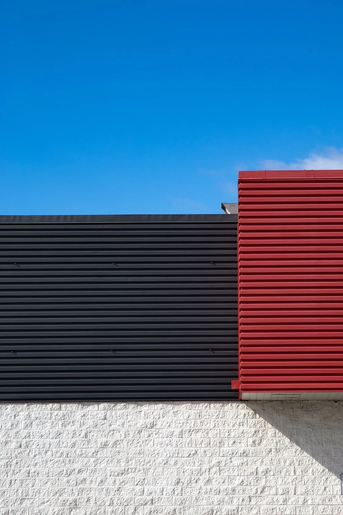 Black and red corrugated metal panels over a white brick wall under blue sky, part of Lionel May's photography series on suburban vernacular architecture.