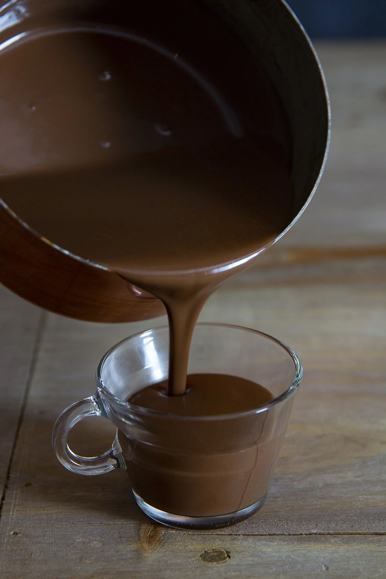Thick dark chocolate being poured from copper pot into a small drinking cup, on rustic wooden table