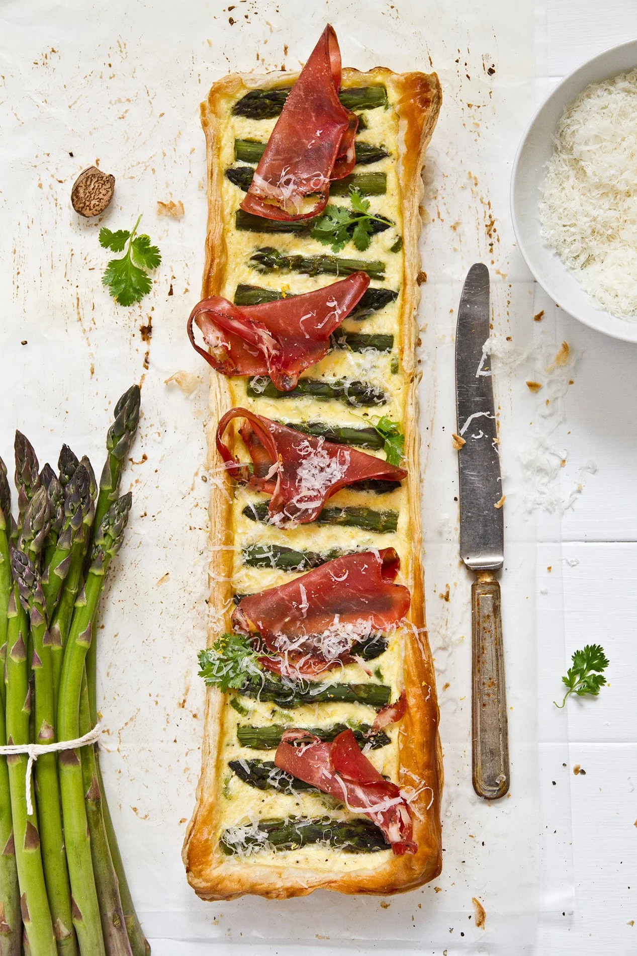 Overhead shot of an asparagus and ricotta tart topped with coppa curls and grated parmesan, flanked by fresh asparagus and a knife.