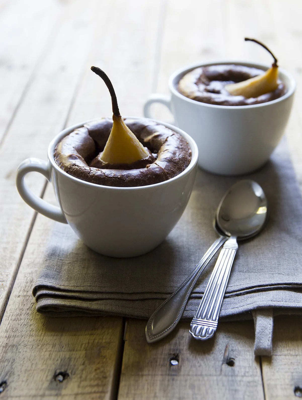 Dark chocolate cakes baked in white cups, with a poached pear directly in the cake, on cloth and wooden table, with silver spoons. 