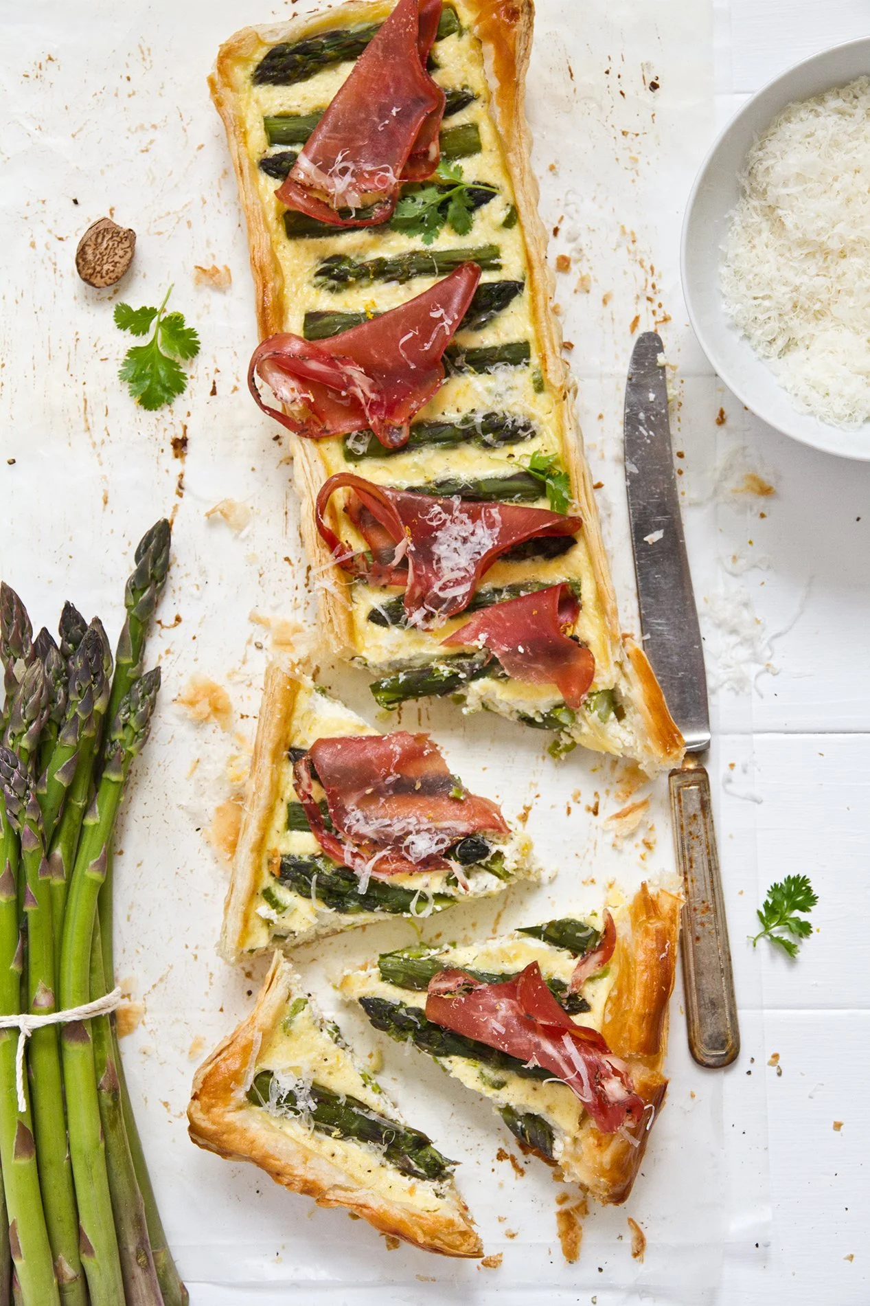 Kitchen photo of a cut asparagus and ricotta tart topped with grilled coppa and grated parmesan flakes with asparagus.