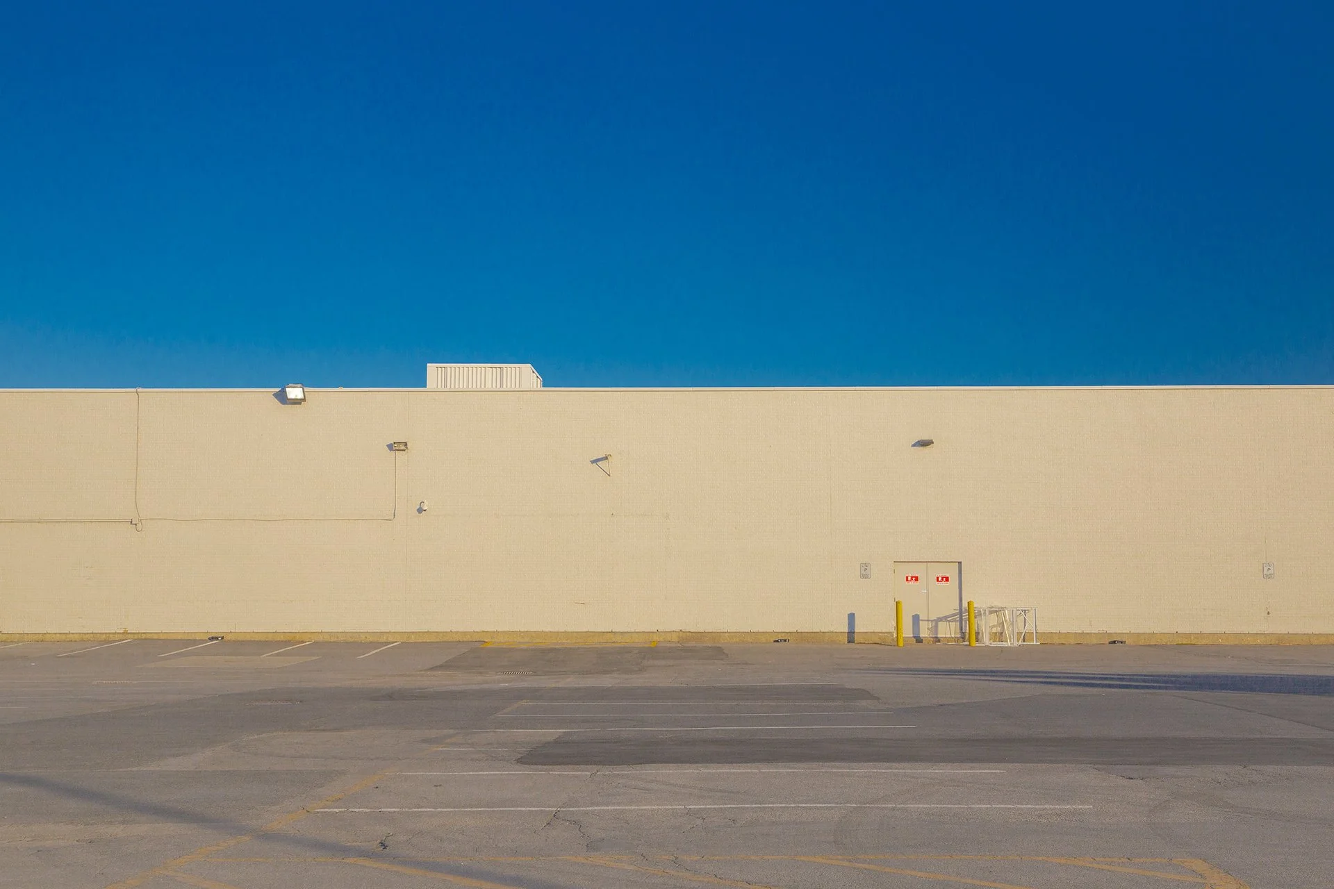 Minimalist horizontal bands of empty parking lot, beige warehouse wall, and clear blue sky.