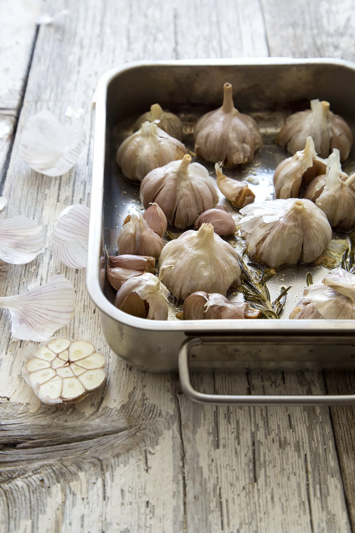 Whole heads of garlic roasting in a metal pan with rosemary and olive oil, loose garlic peels on the wood table.