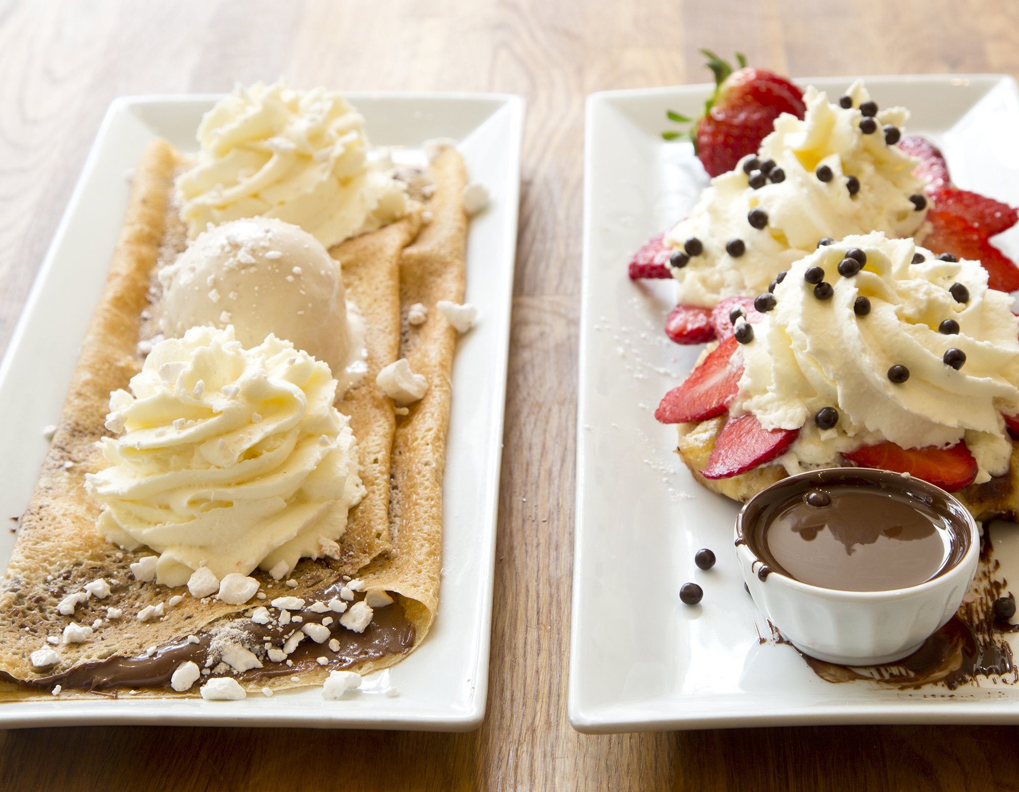 Two gourmet french crepes garnished with hazelnut spread, ice cream, whipped cream, fresh strawberries and melted chocolate on long serving plates