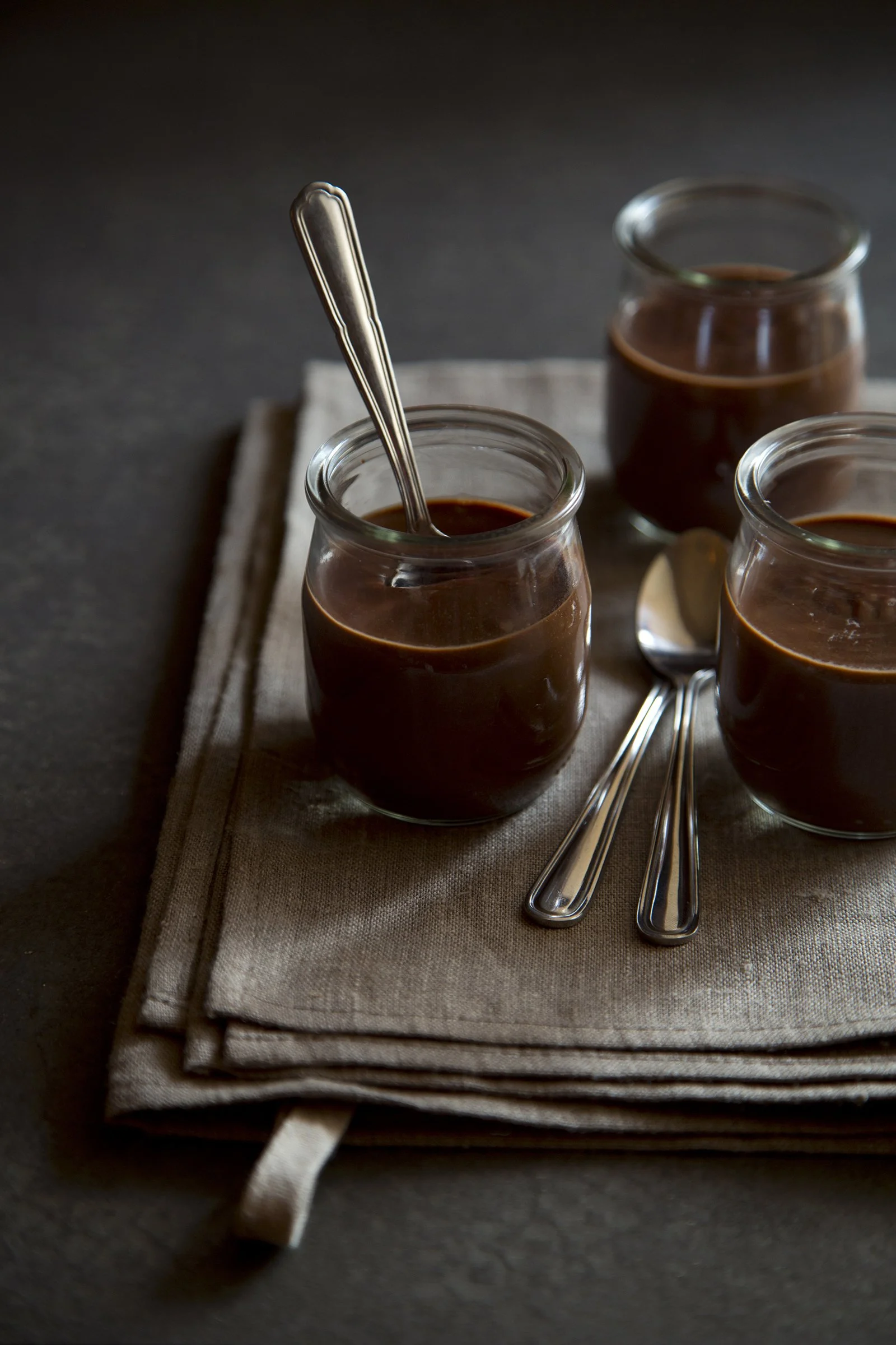 Dark bittersweet chocolat pot de creme, on grey nakpin and dark table, with silver spoon stuck in the chocolate cream