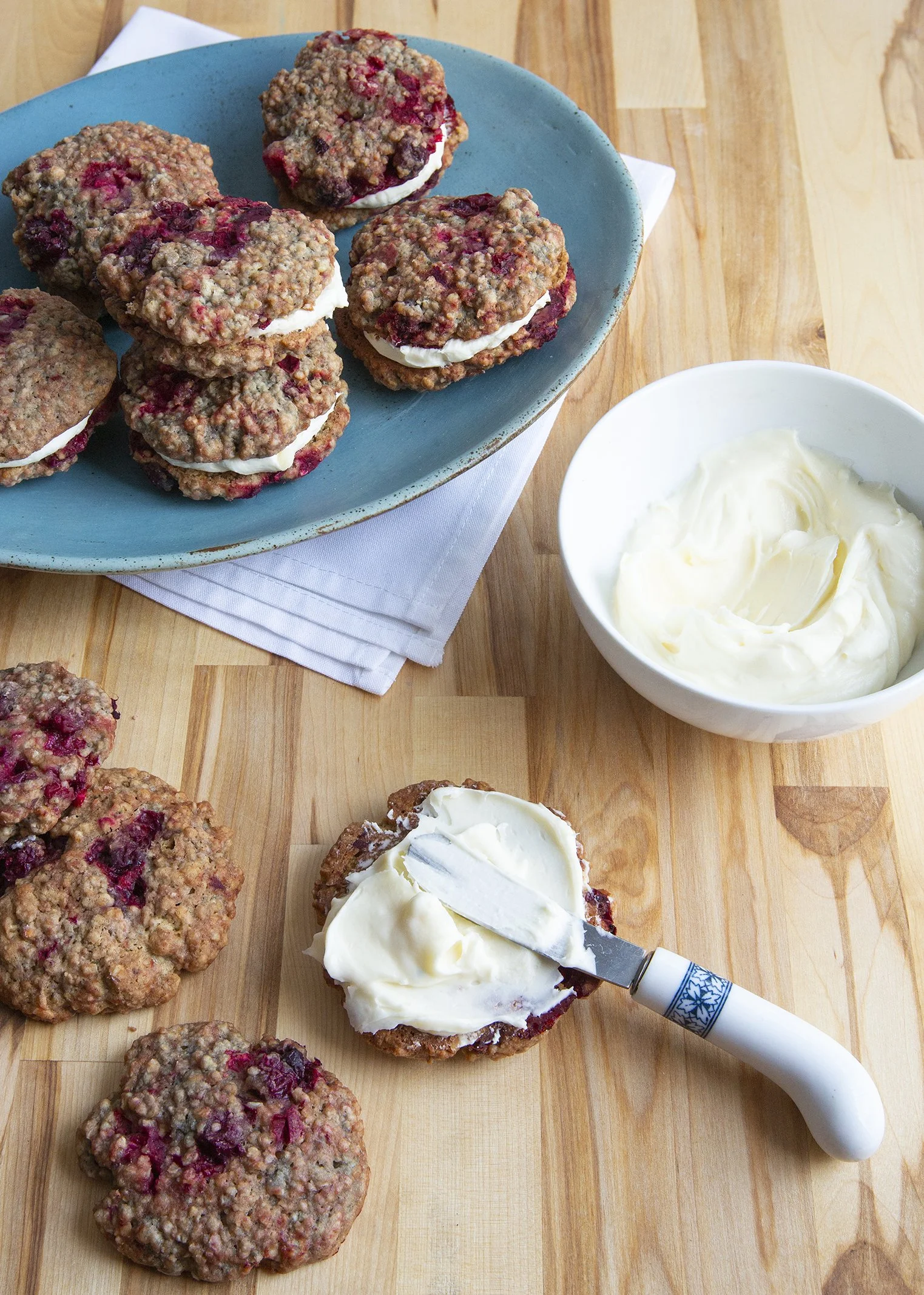 Cranberry cookies with cream cheese interior, kitchen shot with cookies being decorated with bowl of frosting and small knife on butcher block