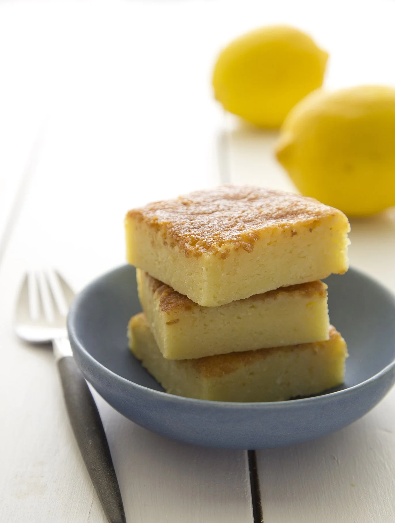 Bright yellow lemon and white chocolate brownies, on blue serving plate on white wooden table, with lemons in the background.