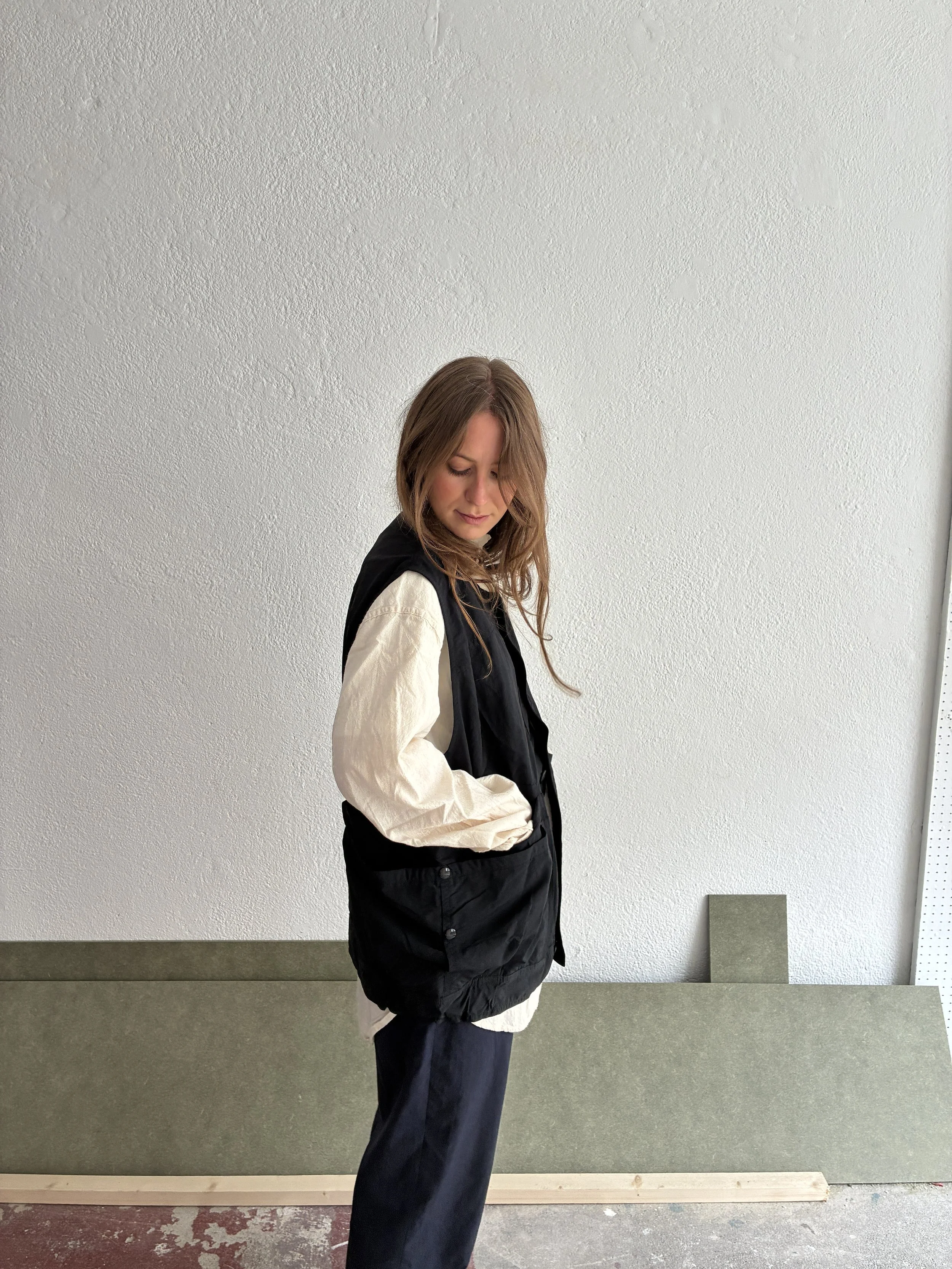 Side profile of Woman wearing Black Sassafras Overgrown Hiker Vest in front of concrete wall