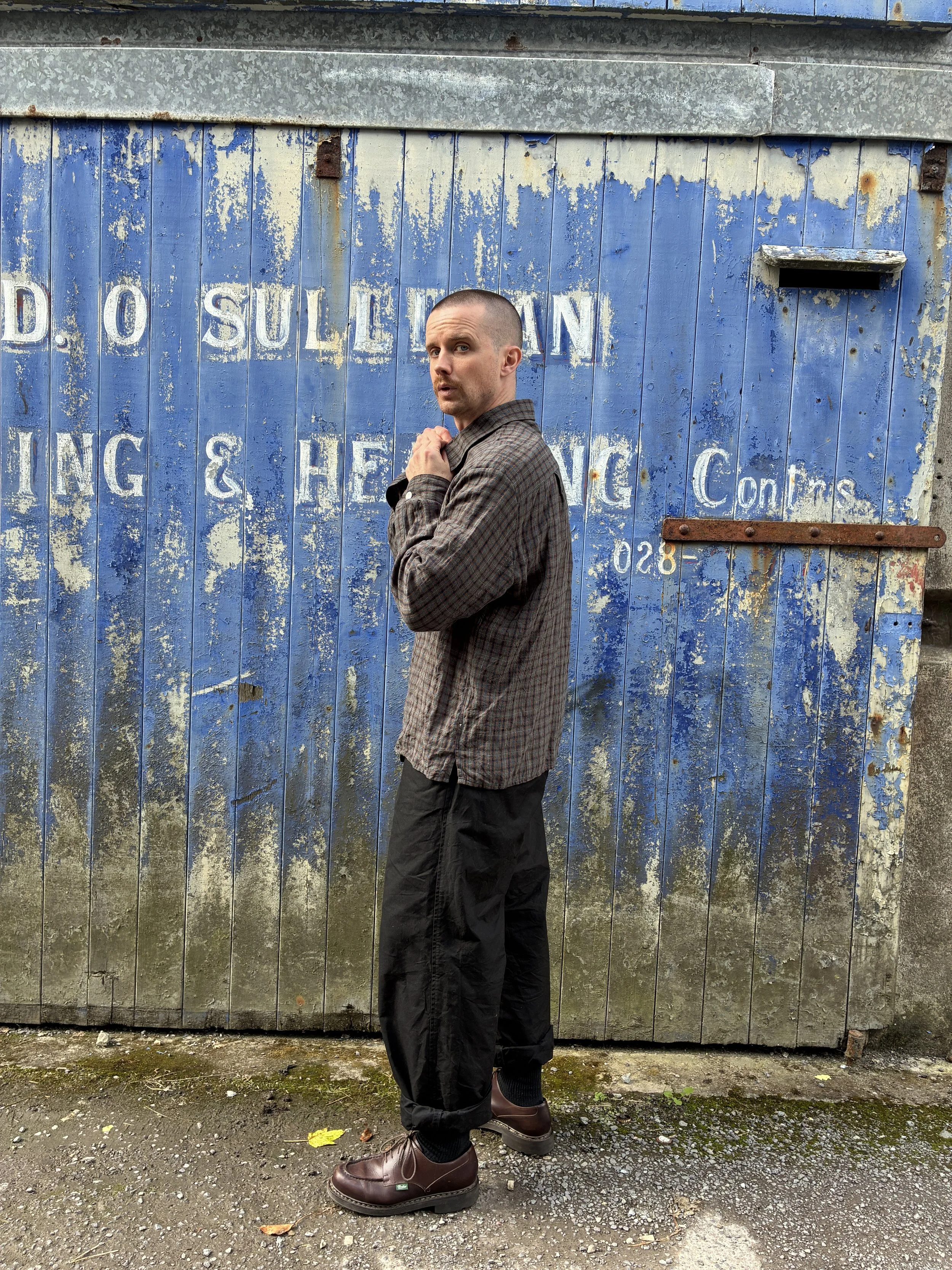side of Man wearing Dusk Conkers Relaxed Farmer Shirt in front of rusty blue shutter