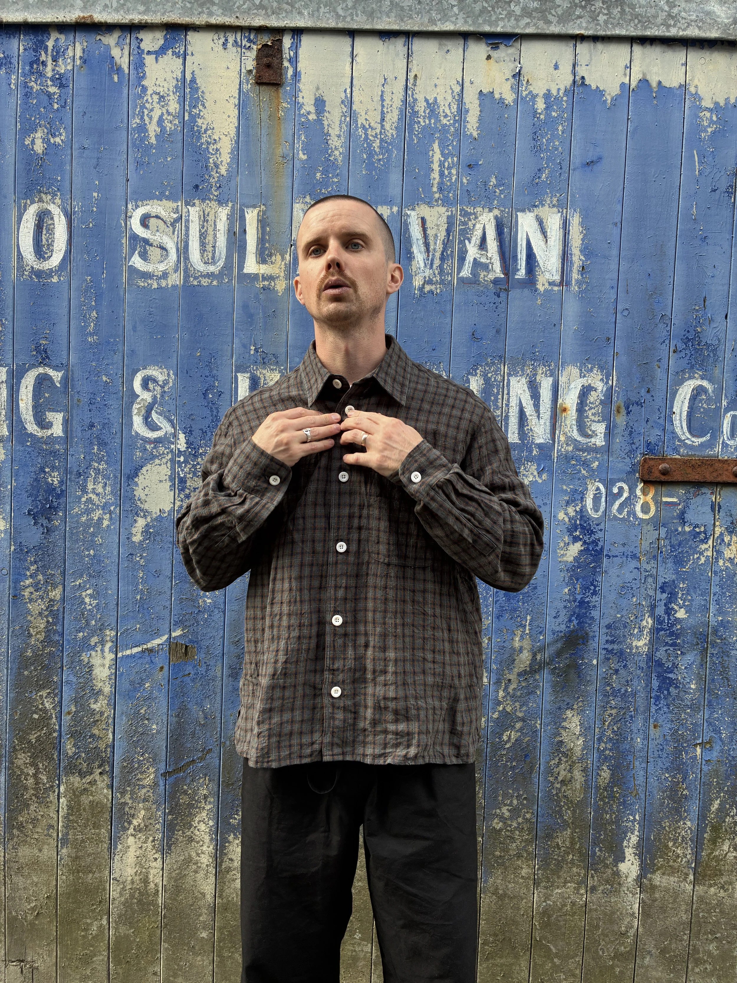 Man buttoning up Dusk Conkers Relaxed Farmer Shirt in front of rusty blue shutter