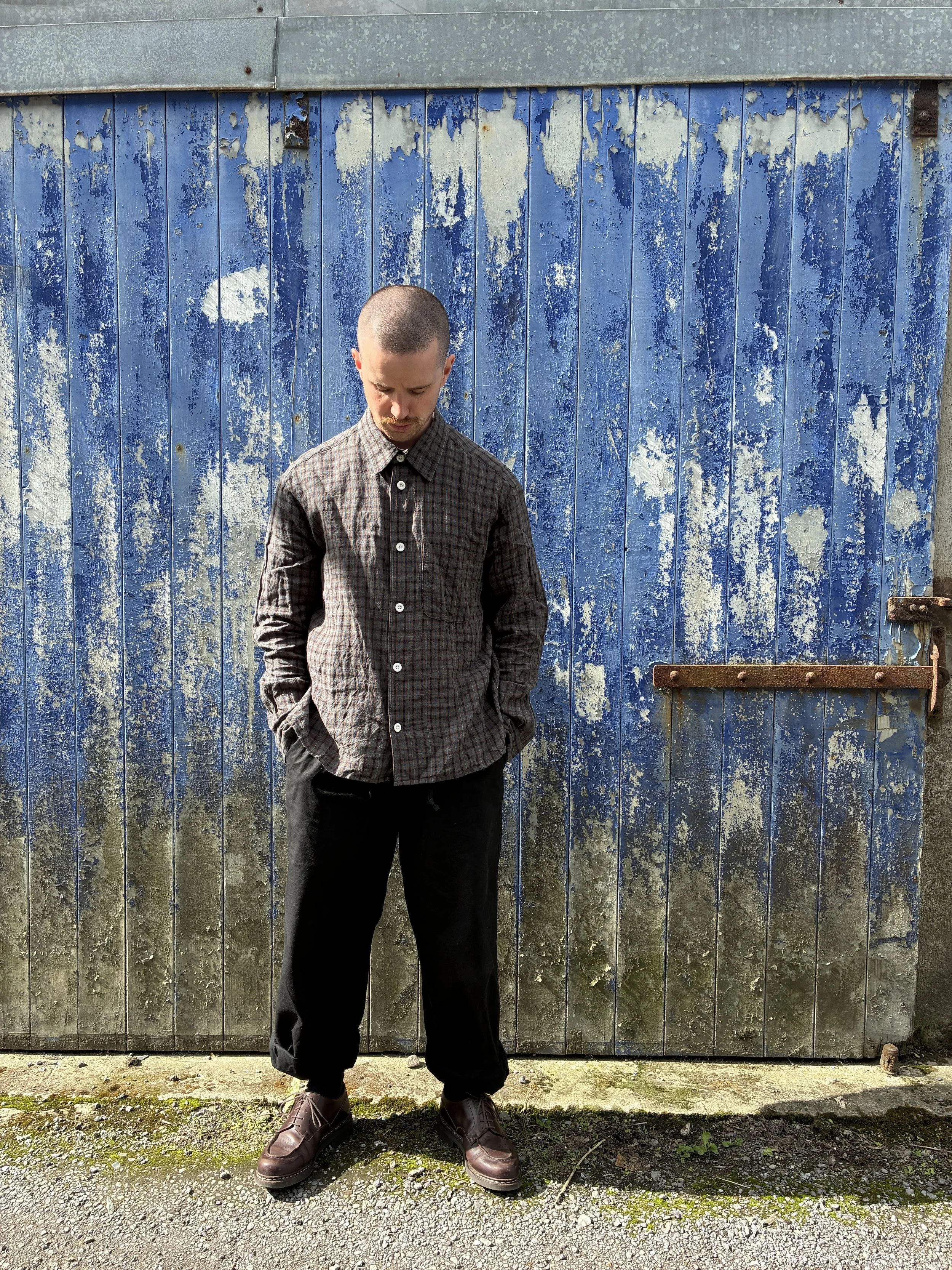 Man wearing Dusk Conkers Relaxed Farmer Shirt in front of rusty blue shutter looking down at ground