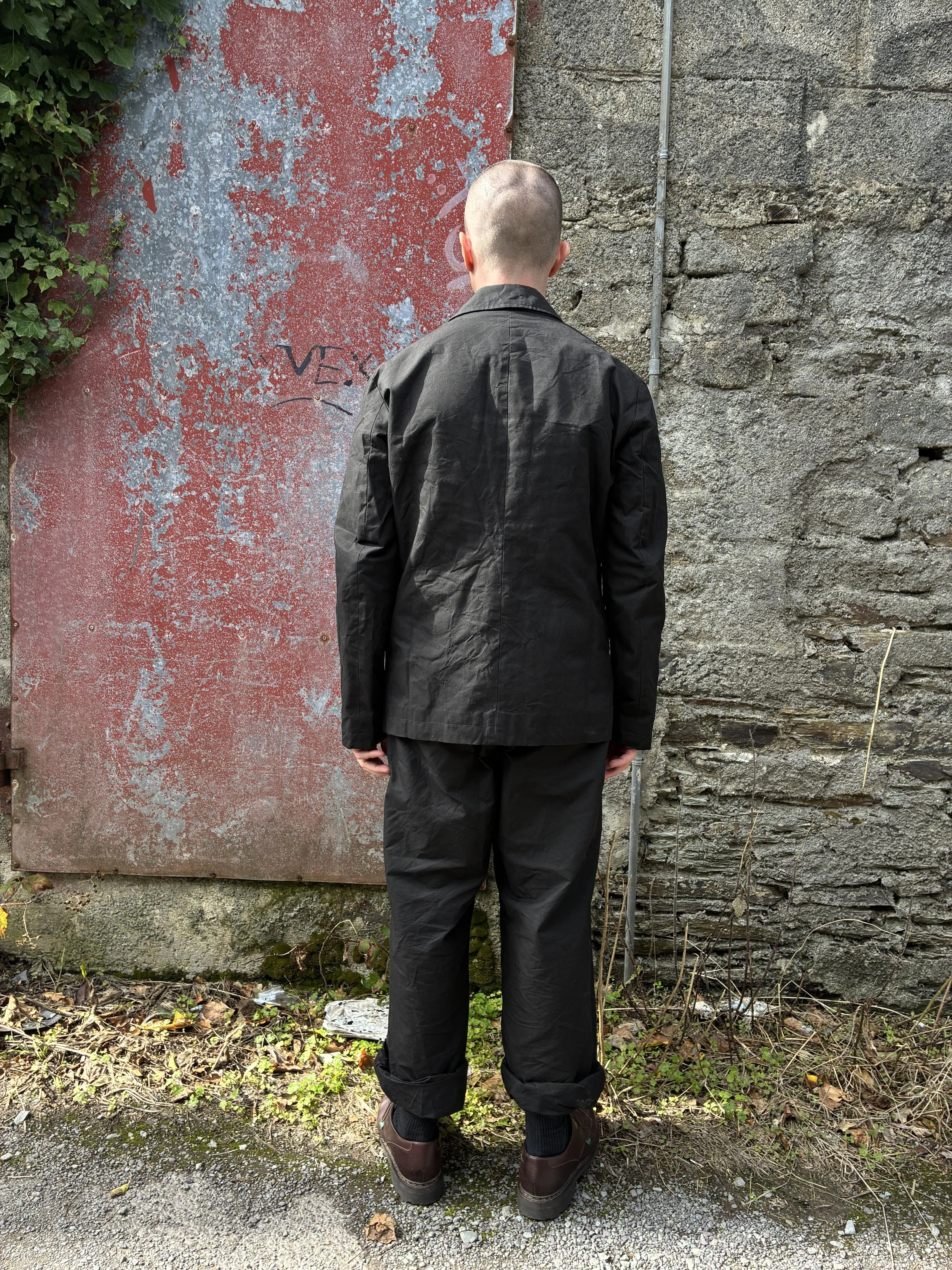 Back of Man in front of red wall outside wearing Black Conkers Rambler Blazer and trousers