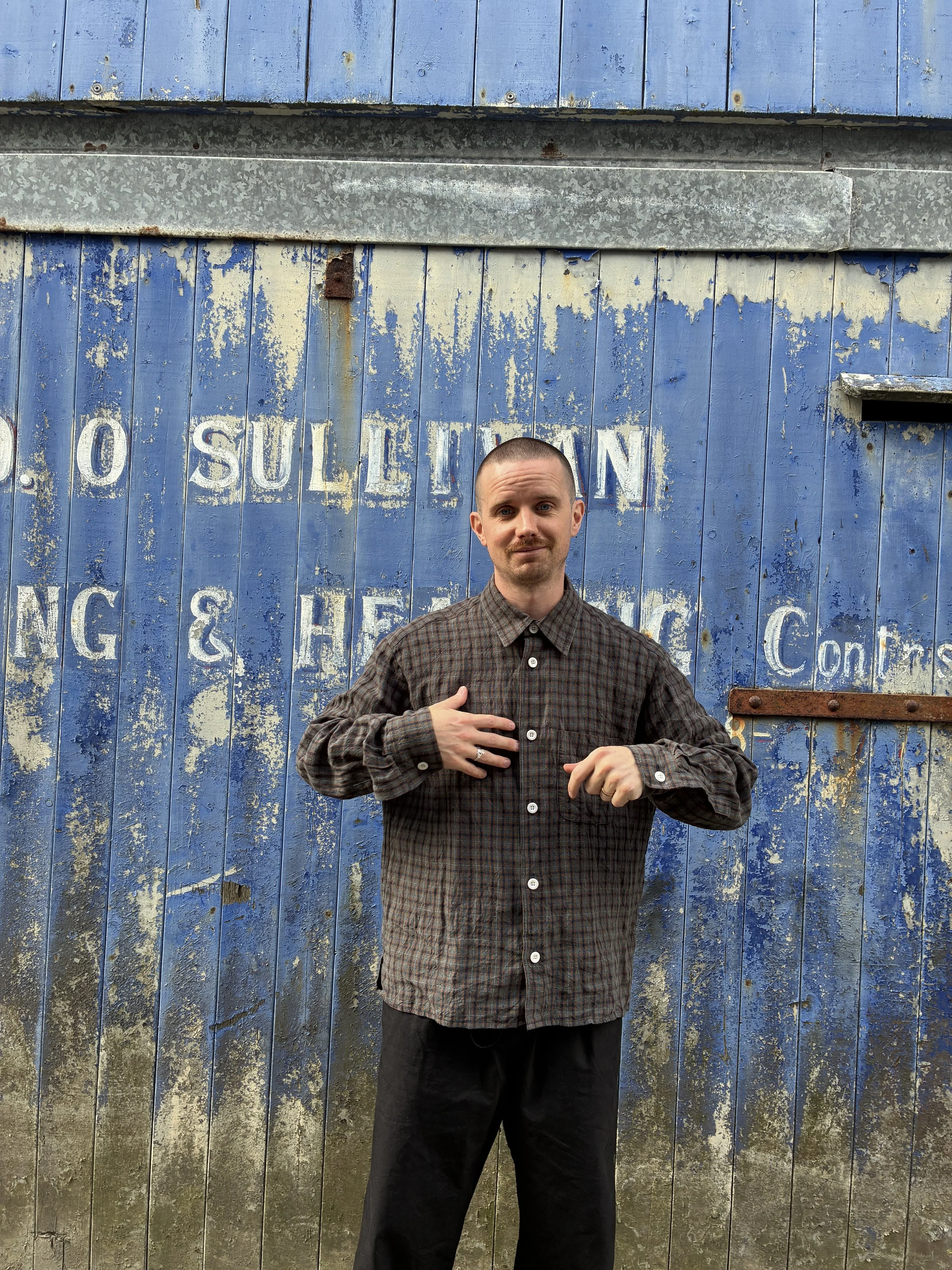 Man wearing Dusk Conkers Relaxed Farmer Shirt in front of rusty blue shutter smiling