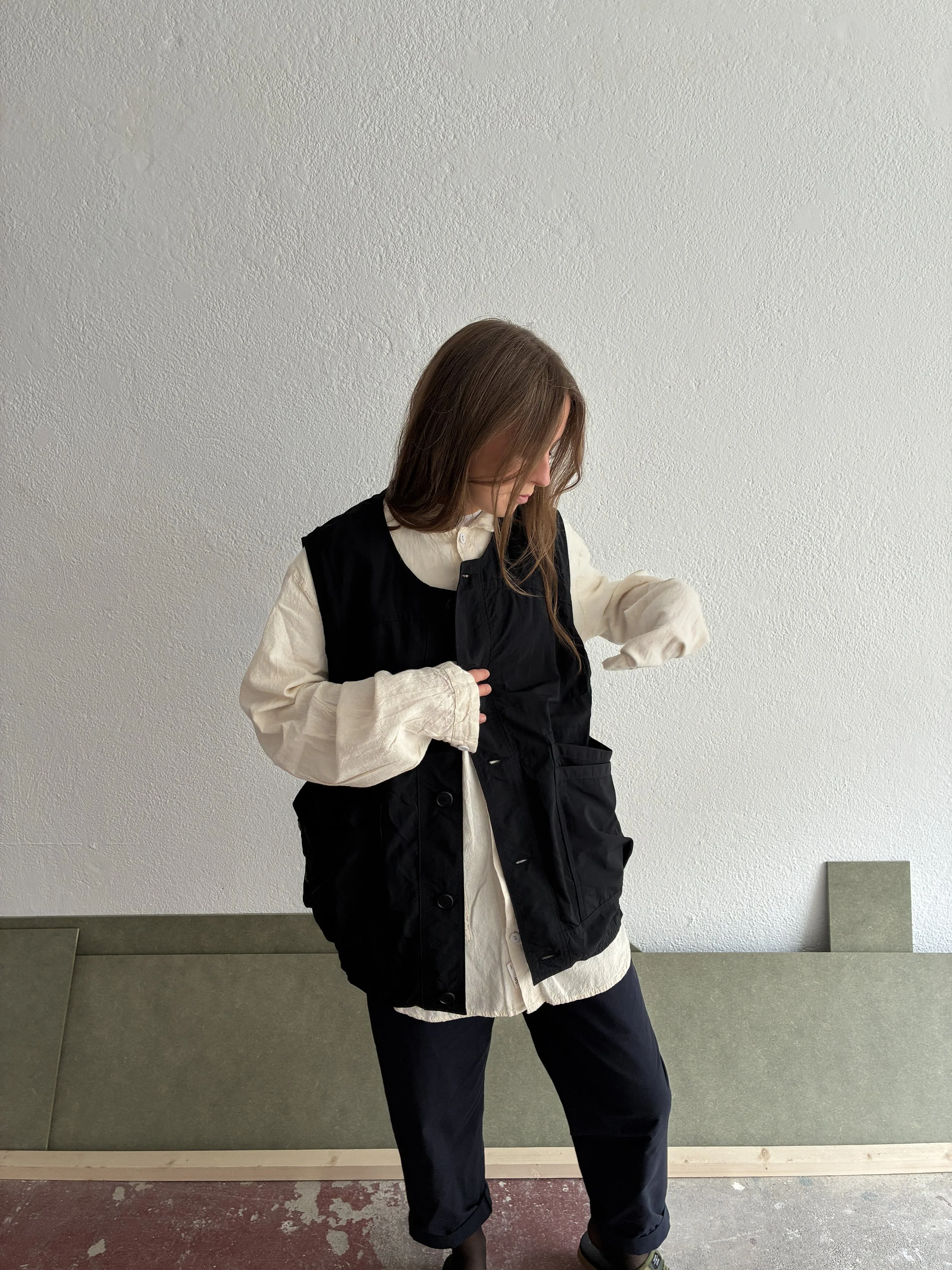Woman looking down wearing Black Sassafras Overgrown Hiker Vest in front of concrete wall