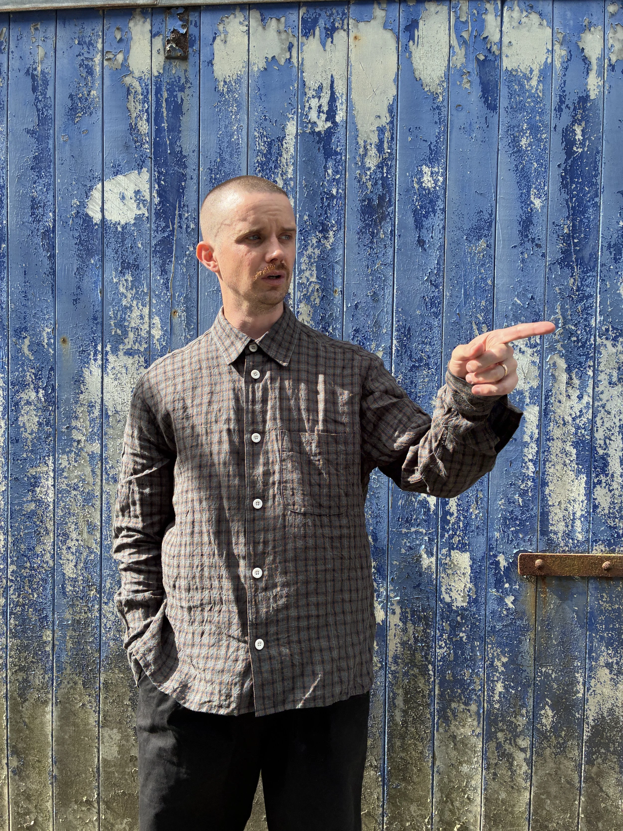 Man wearing Dusk Conkers Relaxed Farmer Shirt in front of rusty blue shutter while pointing