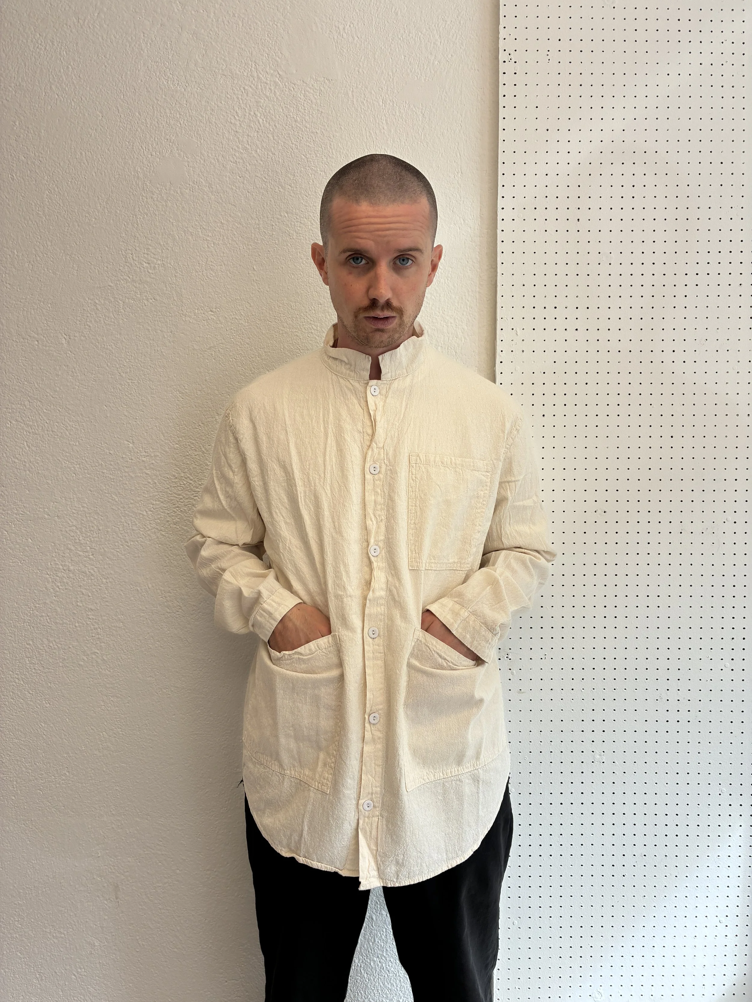 man with hands in pockets of Tender Co Type 457 ecru rinsed Frock Shirt in front of white wall