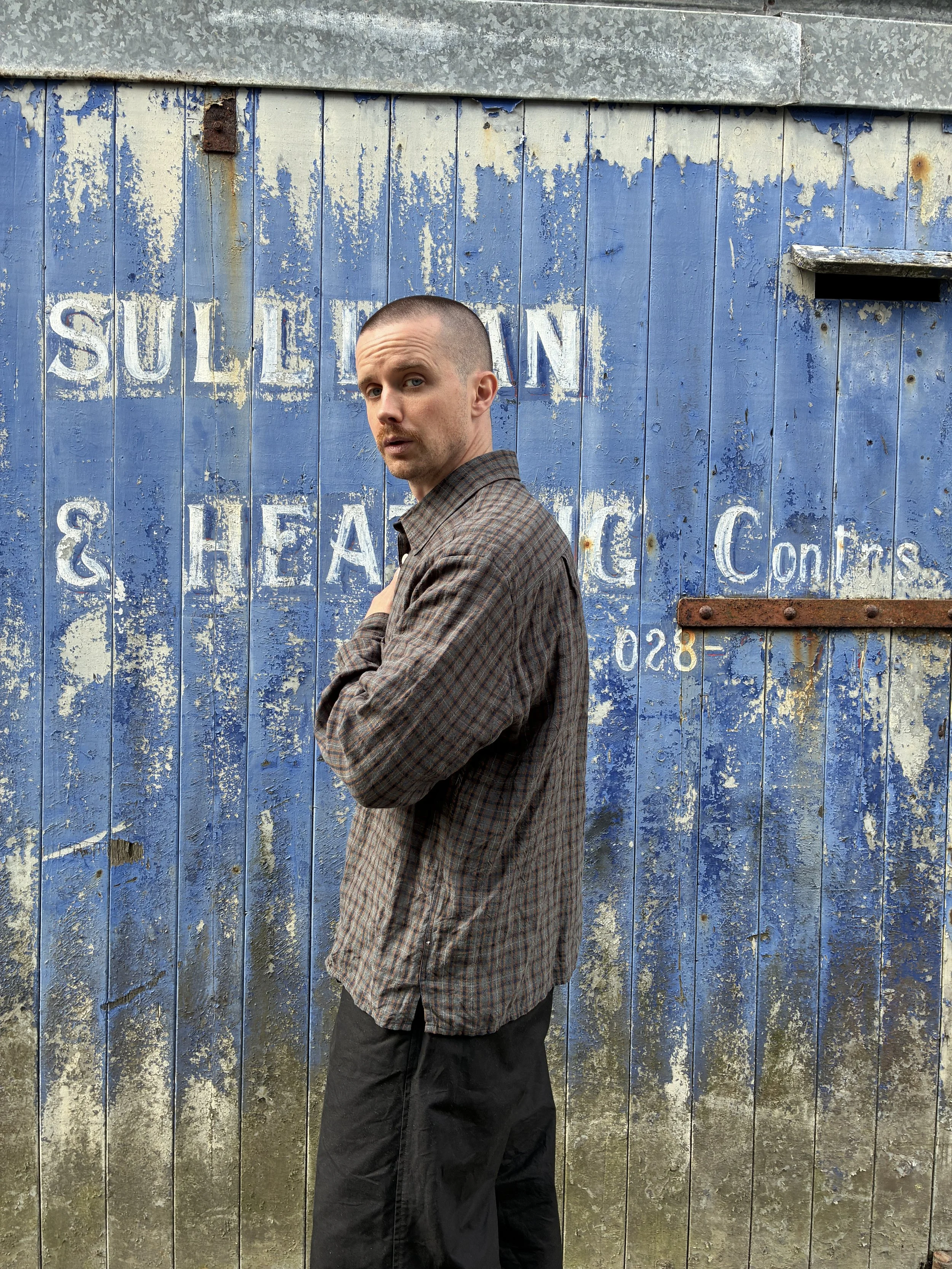 Man wearing Dusk Conkers Relaxed Farmer Shirt in front of rusty blue shutter