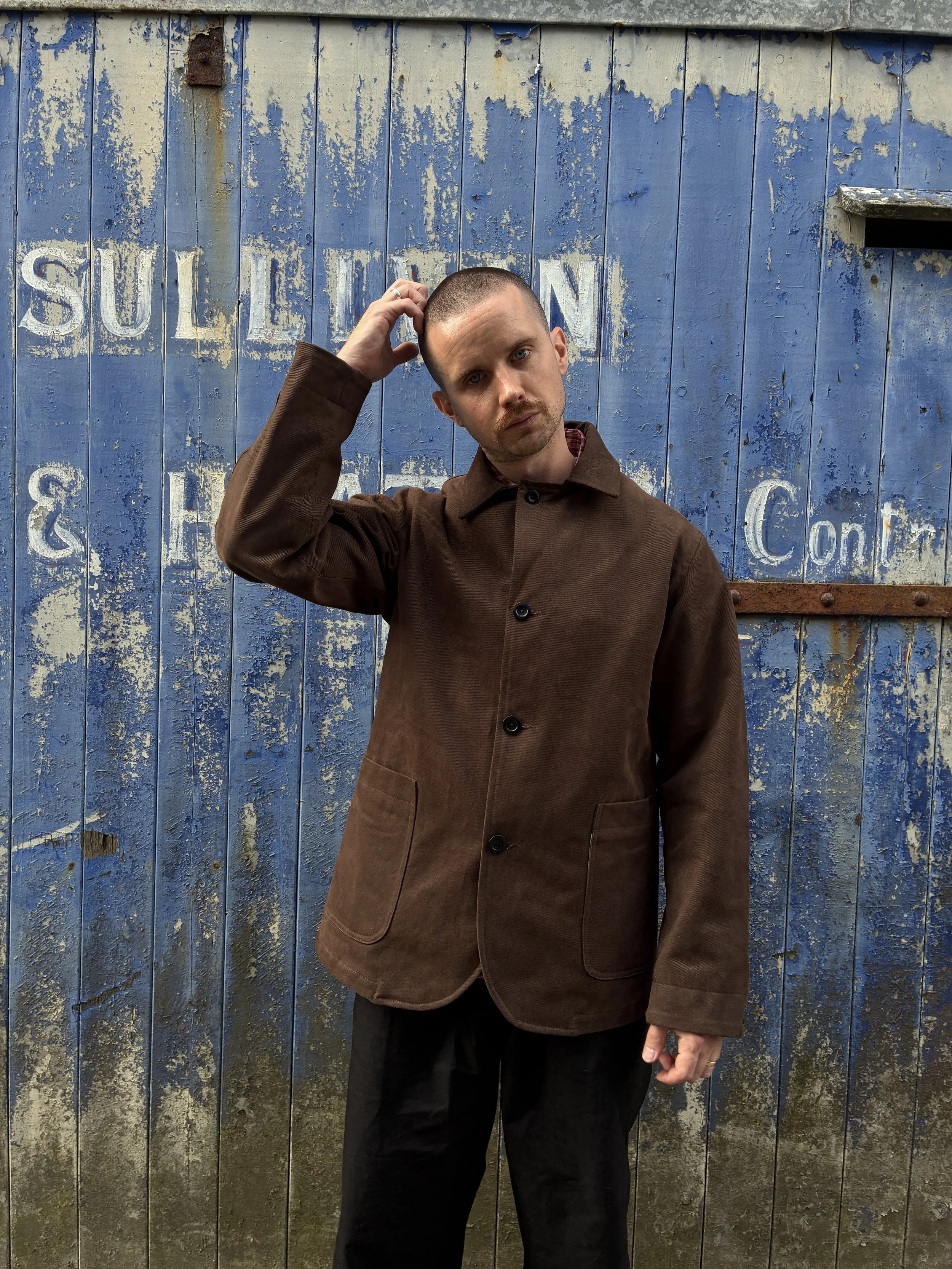 Man in front of blue rusty door wearing brown Conkers Bruton Blazer Jacket while tilting head
