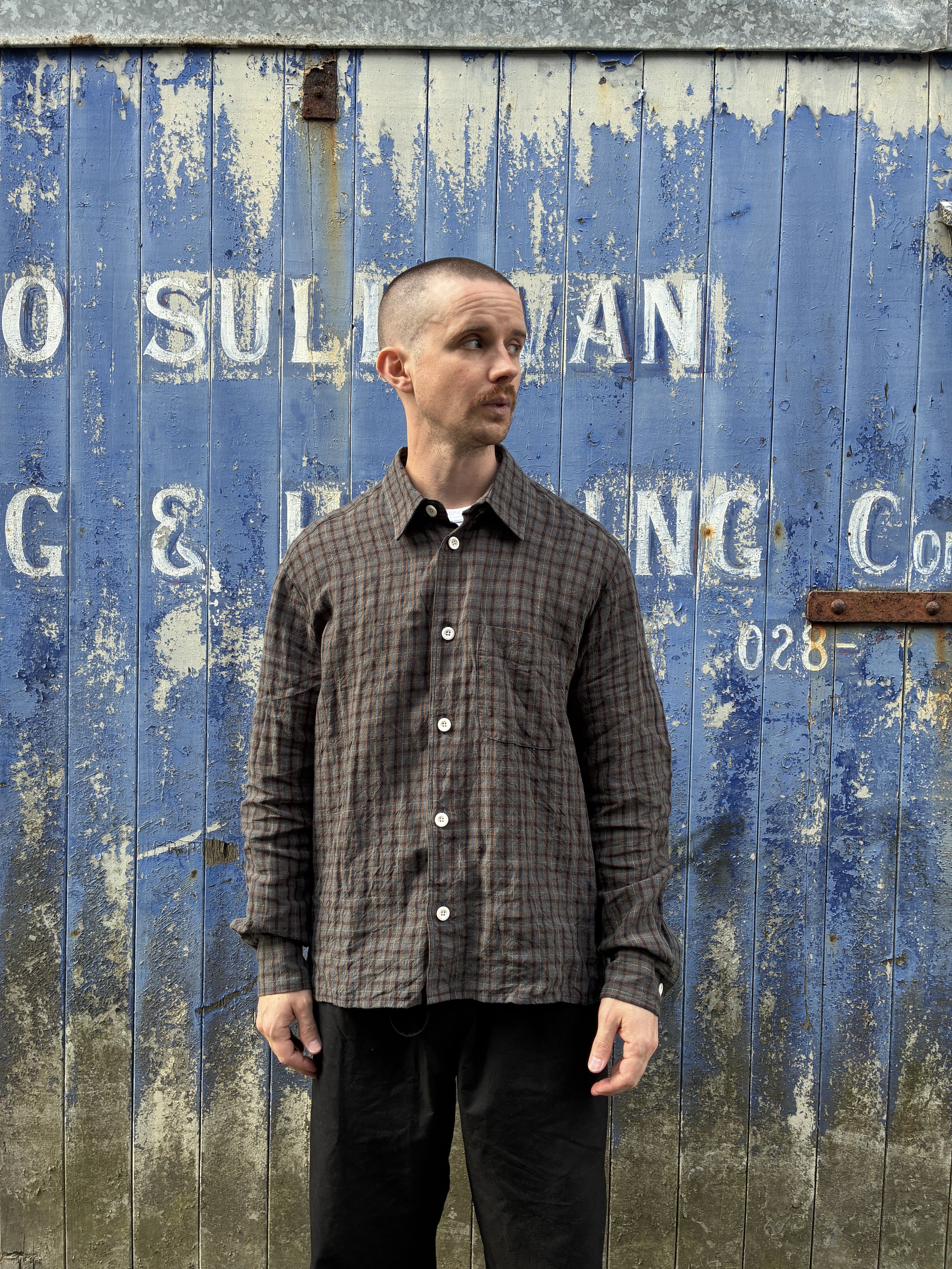 Man wearing Dusk Conkers Relaxed Farmer Shirt in front of rusty blue shutter