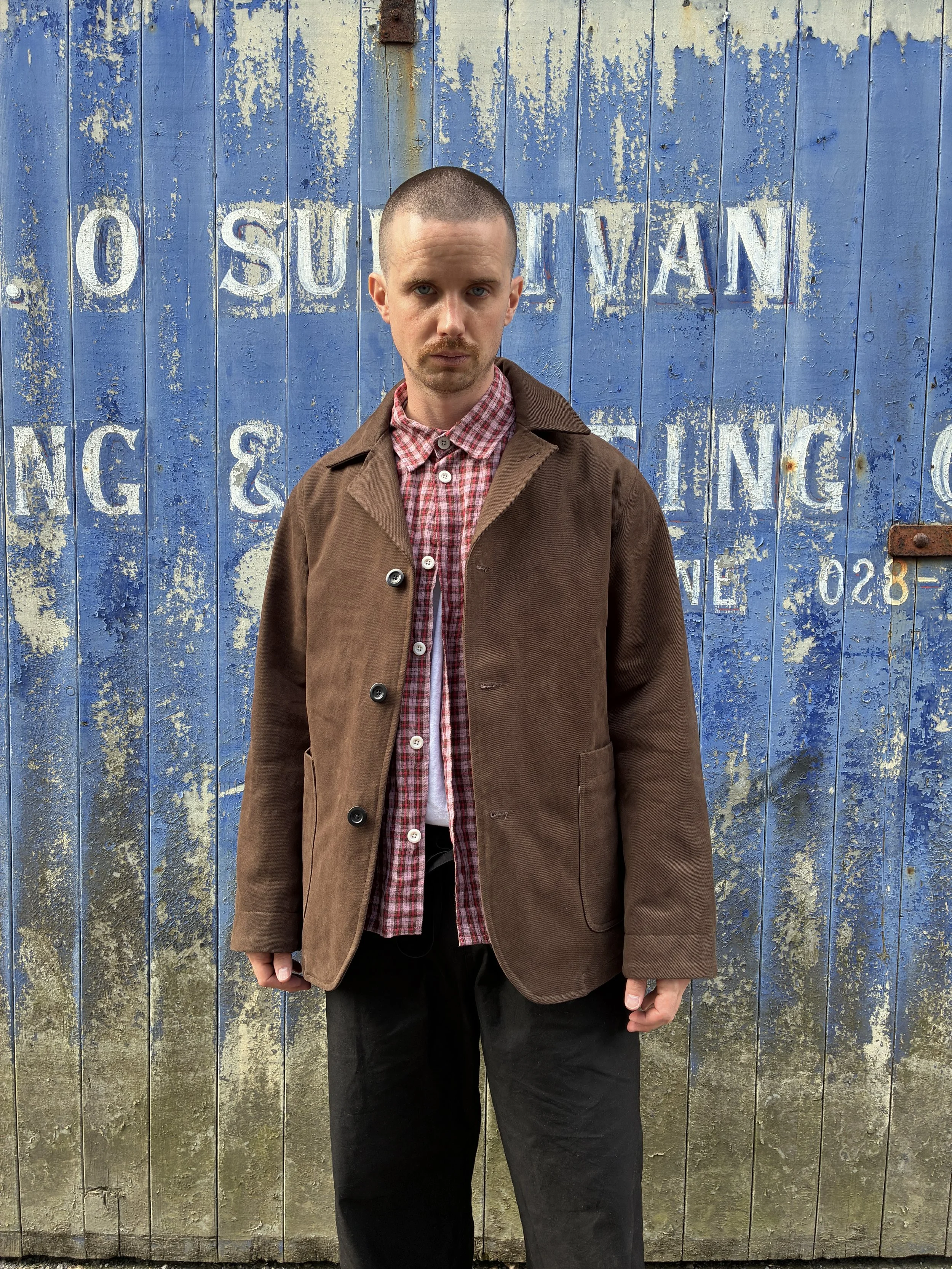 Man in front of blue rusty door wearing brown Conkers Bruton Blazer Jacket