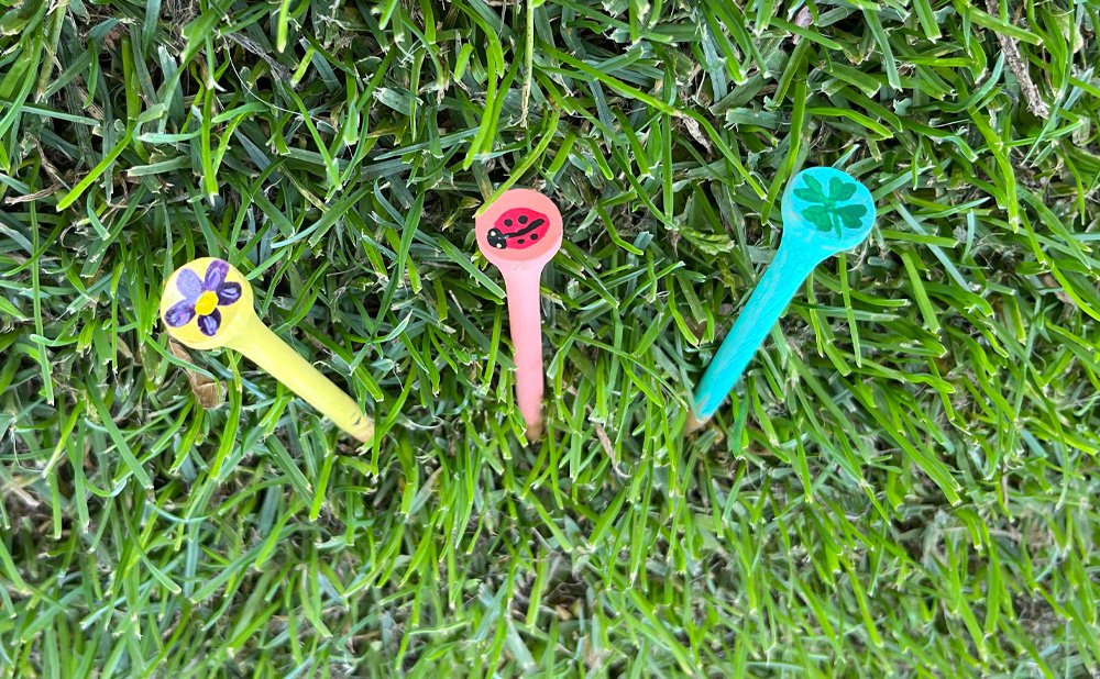 Three golf tees in different summer colors with hand-painted designs on top laying in the grass.