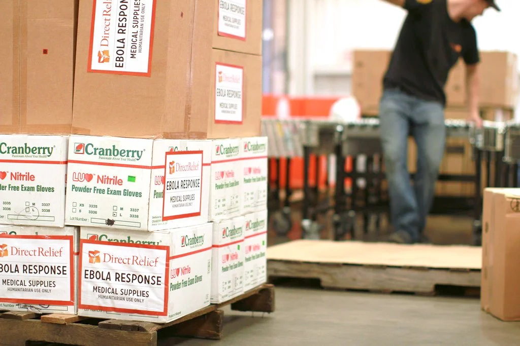 Pallet of boxes labeled "Ebola Response Medical Supplies," including nitrile gloves, in a warehouse setting with a person in the background.
