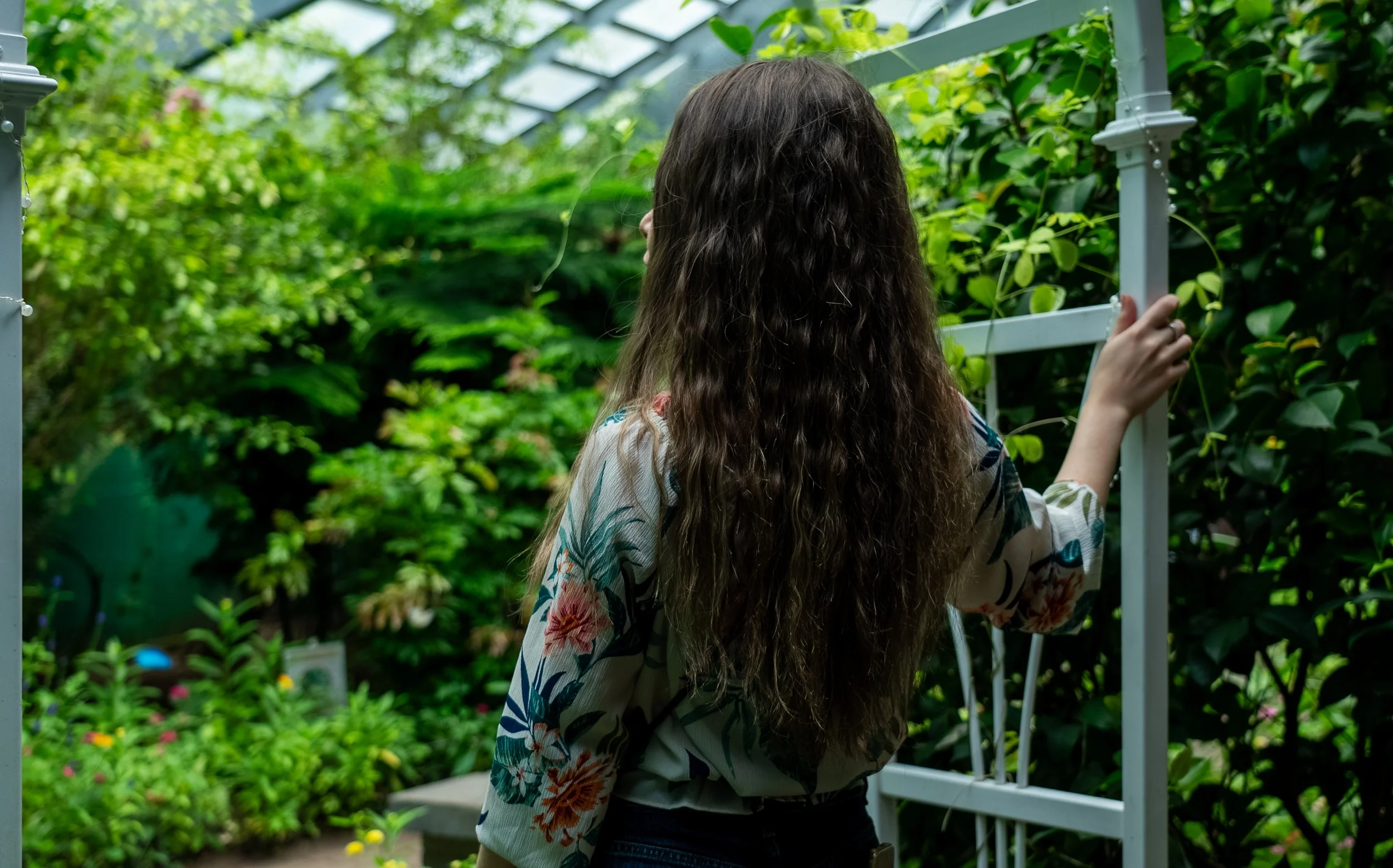 A person with long, curly hair wearing a floral-patterned shirt standing in a lush green garden, facing away and holding a white metal frame.