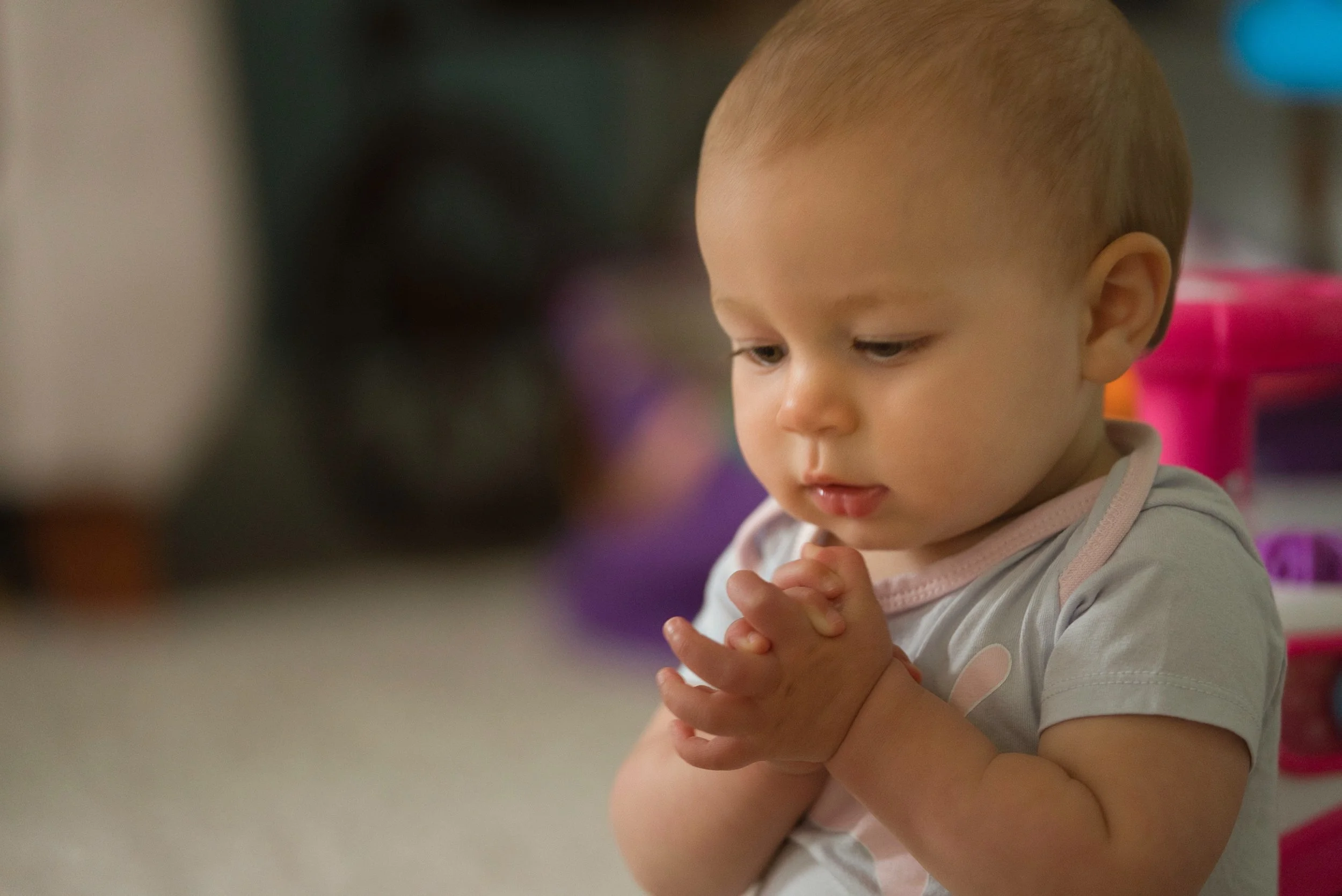 A young child with light skin and short hair is praying, with hands clasped together. The child appears focused and kneels on a carpeted floor in a home or daycare setting, with colorful toys visible in the background.